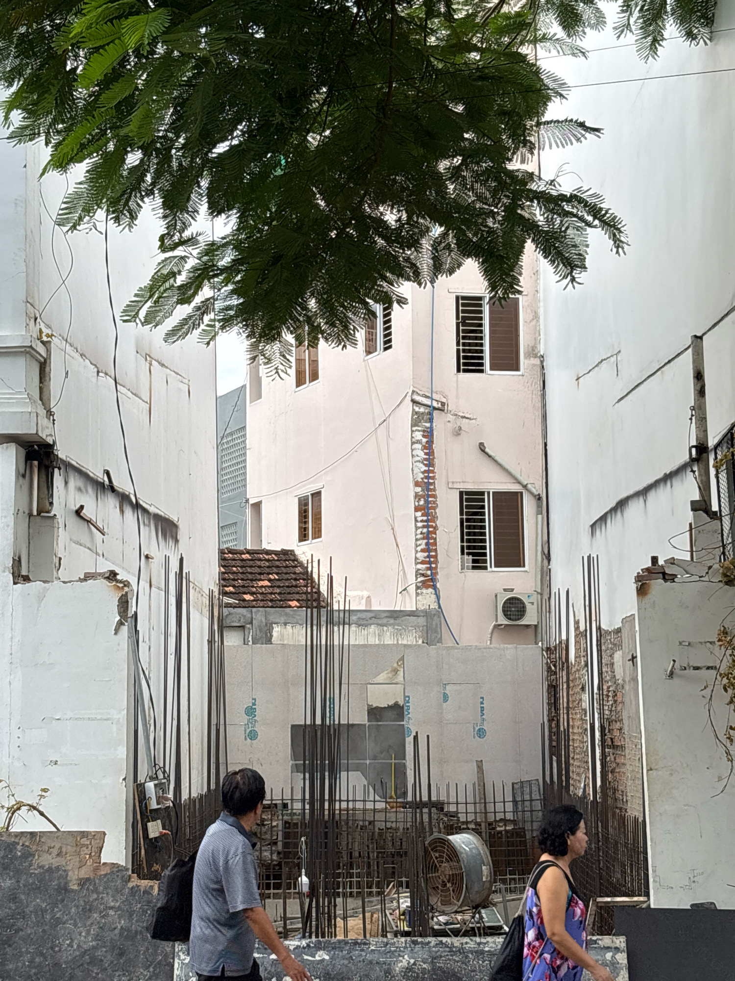 In Nha Trang, Vietnam, a narrow street is depicted with buildings on either side. A large tree with dark green foliage overhangs the scene from the top. The building on the left is white with exposed wiring and a damaged facade. The building on the right is light pink with multiple windows, some with louvers. In the background, a multi-story building with a grid-like facade is visible. The foreground shows a construction site with rebar, concrete panels, and a large industrial fan. Two individuals are walking past the construction site. The person on the left is a man wearing a striped shirt and dark pants, carrying a black backpack. The person on the right is a woman wearing a floral-print dress and carrying a black bag over her shoulder. The scene appears to be under construction or renovation, with materials and scaffolding present.