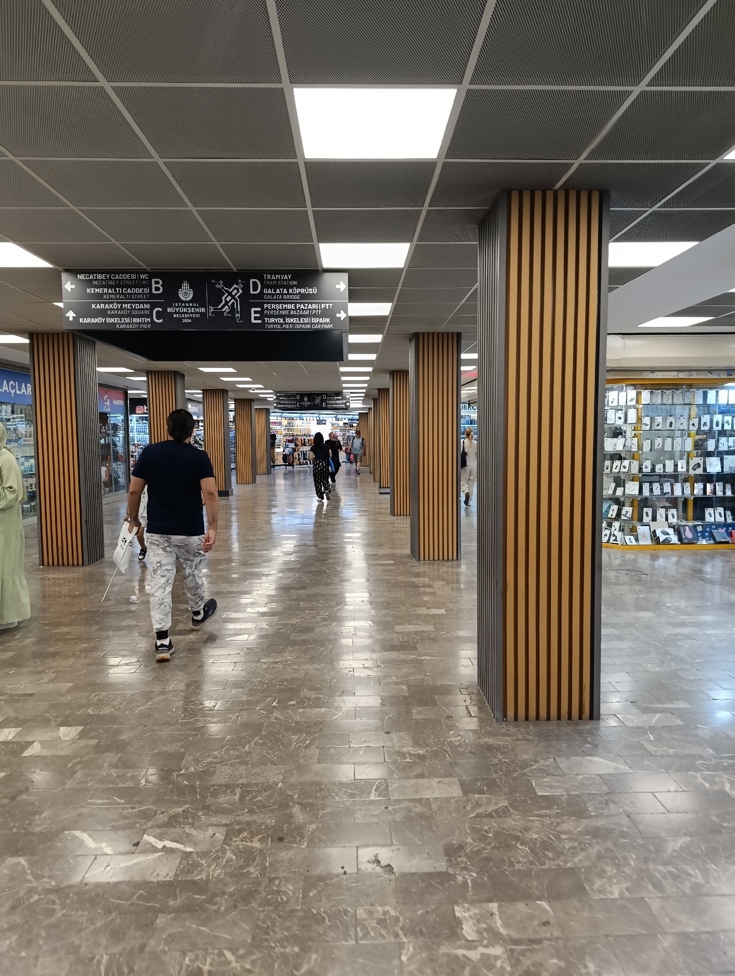 An interior shot captures a man in camouflage pants and a dark t-shirt walking away from the camera down a well-lit, marble-floored corridor. The corridor is lined with columns clad in vertical wooden slats, and fluorescent lights illuminate the ceiling. To the left, a storefront is partially visible, and to the right, a display of various electronic accessories is showcased in a glass cabinet. Overhead signs provide directional information for "NECATIBEY CADDESİ WC," "KENERALTI STREET," "KARAKÖY MEYDANI," "KARAKÖY SQUARE," "KARAKÖY ISKELESI," "TRAMVAY," "GALATA KÖPRÜSÜ," "PERSEMBE PAZARI," and "TURKOL PIER / ISPARK CAR PARK." The setting appears to be a transportation hub or a commercial area within Istanbul's Beyoğlu district, a vibrant and historic part of the city known for its cultural attractions and bustling atmosphere. Several other people are visible in the background, walking in the same direction as the main subject, suggesting a public space with ongoing activity.