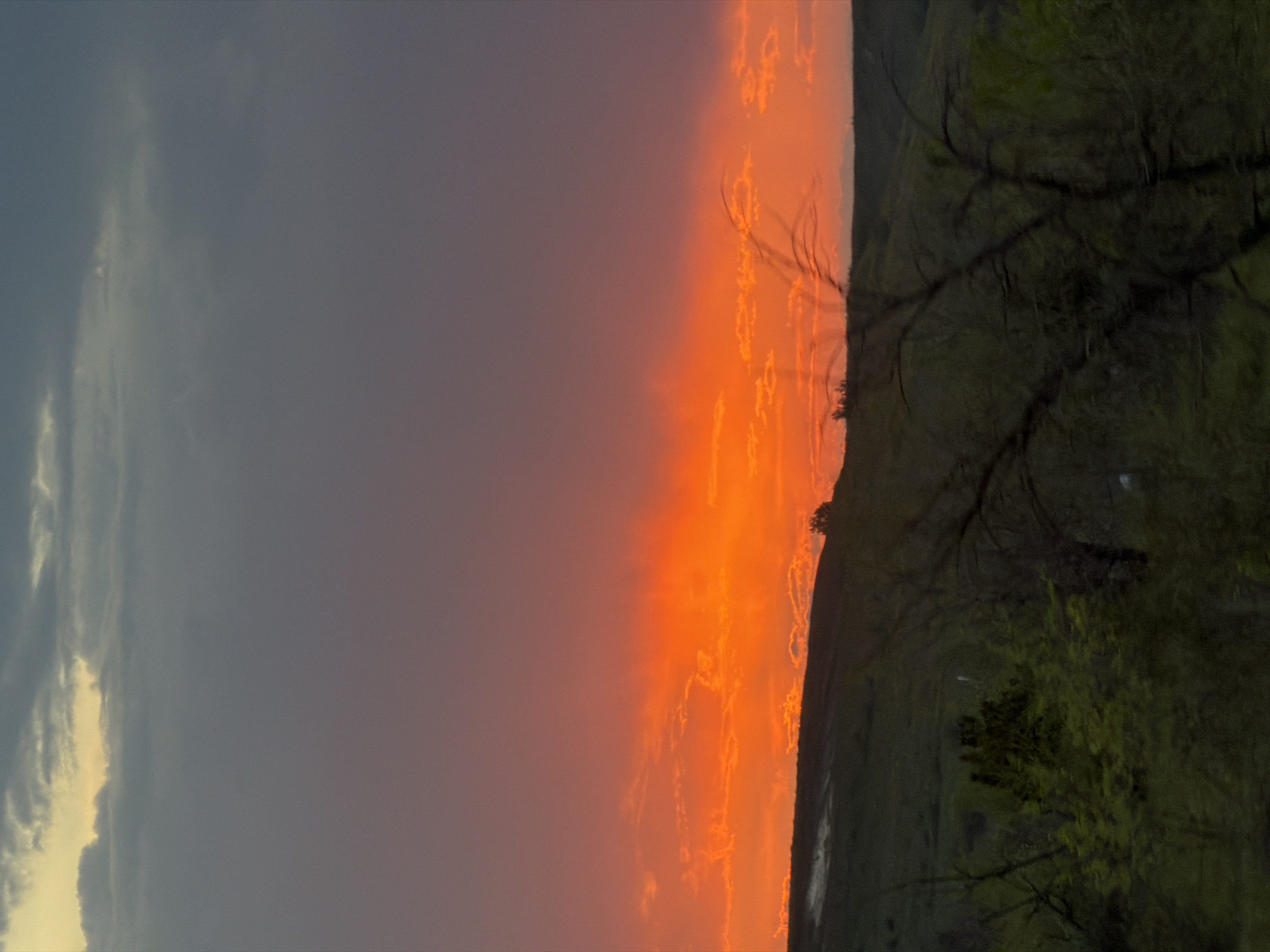 The image shows a landscape at sunset. To the left is a dark, cloudy sky with some light breaking through the clouds. In the center, the sky transitions to a bright orange hue, likely from the setting sun. To the right is a dark landmass with trees and vegetation silhouetted against the bright sky. The location is identified as Ternovka, Russia, though specific features confirming this are not visible in the image.