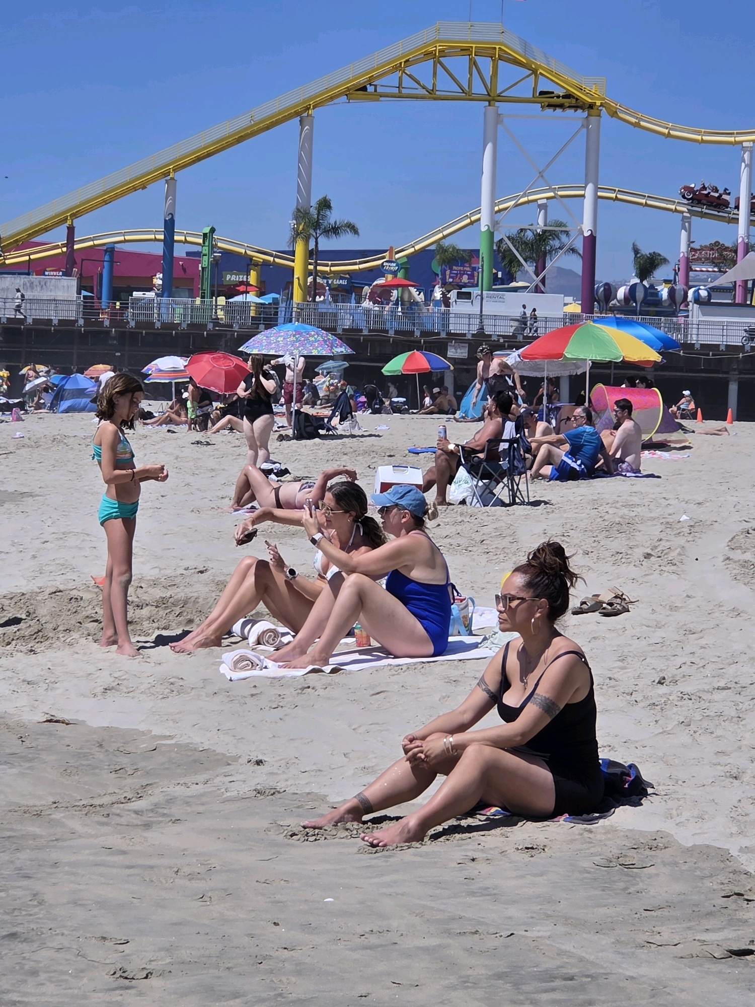 The image depicts a lively beach scene in Santa Monica, California, under a bright, sunny sky, suggesting it's midday. A roller coaster, a prominent amusement park feature, looms in the background, its yellow tracks curving against the clear blue sky. In the foreground, several people are relaxing on the sand, some lying on towels and others sitting. A young girl in a turquoise bikini stands near the left side of the frame, appearing to interact with someone out of view.
Umbrellas of various colors, including red, blue, and a colorful rainbow-striped one, are scattered across the beach, providing shade for beachgoers. People are dressed in swimwear, indicating a typical beach day activity. Some are seen sunbathing, while others appear to be engaged in conversation or simply enjoying the atmosphere. A woman in a black swimsuit sits in the foreground, her arms crossed and looking to the side, possibly in contemplation or observing her surroundings. Her arms are adorned with tattoos. In the mid-ground, other individuals are also visible, some sitting under umbrellas, highlighting the social and recreational nature of the setting.
The presence of the roller coaster and the general ambiance of people enjoying a sunny day on the beach strongly suggests a location associated with an amusement pier or boardwalk, such as the Santa Monica Pier. The overall mood is relaxed and summery, with the vibrant colors of the umbrellas and the roller coaster contributing to a cheerful atmosphere. No specific text is clearly legible in the image, other than what appears to be some signage on the pier structures in the background.