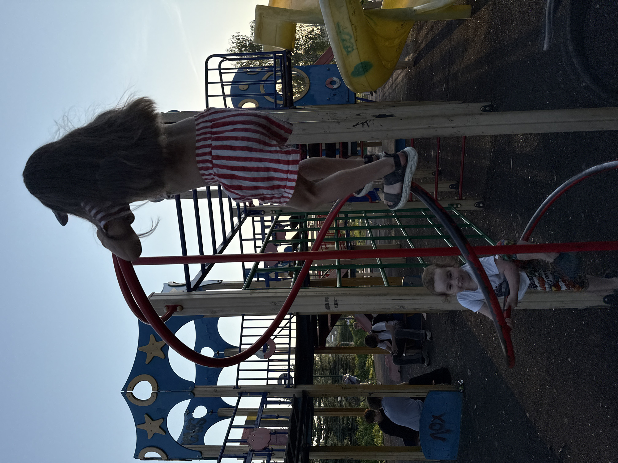 The image shows a playground. A girl with long brown hair and a red and white striped top and shorts is hanging upside down from a red metal bar. She is wearing sandals. Another younger child in a white t-shirt and patterned shorts is holding onto a similar red metal bar on the ground, looking towards the camera. The playground equipment is multi-colored and includes ladders, platforms, and slides. Other people are visible in the background, seemingly relaxing on benches under some trees. The sky is visible in the background. The city of Фоки is not discernible.