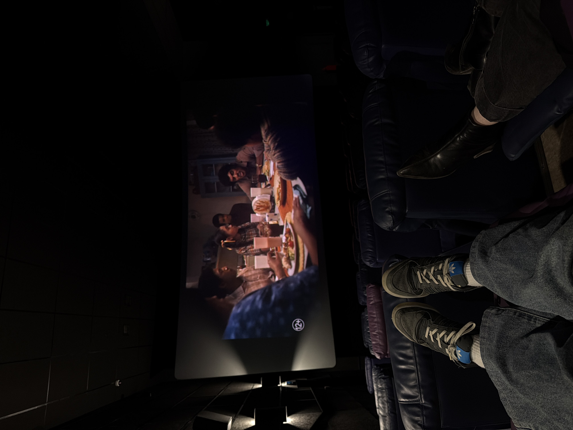 A dim, interior shot captures the experience of watching a movie in a theatre. The angle is from the perspective of someone seated, with their legs and feet in view at the bottom right. The person is wearing jeans and grey Adidas sneakers with white laces. To the right of their legs are dark blue upholstered seats. Further to the right, another individual's legs are visible; this person is wearing dark jeans and black boots, and there is a dark jacket draped over the seat.
The movie screen dominates the center, displaying a film scene featuring a group of people gathered around a table with food. The screen is illuminated, contrasting with the darkness surrounding it. The theater's floor has a geometric pattern illuminated by small recessed lights. The scene lacks contextual information pinpointing the location within Penza, Russia.