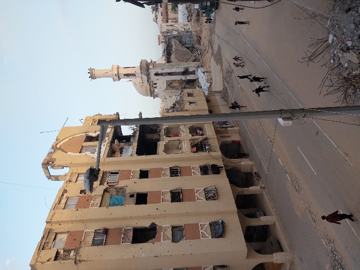 The image displays a devastating scene of urban destruction, likely taken from an elevated position looking down onto a street and surrounding buildings. The photograph is oriented vertically, rotated 90 degrees clockwise, making the street appear as a diagonal path across the frame.
In the foreground and running along the left side of the image is a heavily damaged multi-story building, light brown or beige in color. Large sections of its facade are missing, revealing exposed interiors, broken windows, and debris hanging from balconies and window frames. The upper floors show significant structural compromise, with an entire corner of the building appearing to have collapsed. Pieces of rebar and other structural elements are visible.
Below this damaged building and extending into the mid-ground is a paved road that runs diagonally from the lower right to the upper left. The road itself shows signs of wear and damage, with patches of dirt and debris scattered across its surface. On this road, multiple individuals are visible. A striking detail is the presence of several people lying motionless on the asphalt; some appear to be face up, others face down. Their dark clothing contrasts with the lighter tone of the road. There are at least seven to eight such figures clustered along the street. One individual, seen in the bottom right corner, is standing and appears to be walking away, wearing a red shirt. Another figure further up the street, also in red, might be standing or moving.
In the background, positioned to the right of the street, stands a mosque with a distinctive white minaret and a dome. This structure also appears damaged, with visible signs of destruction around its base and potentially parts of its main building. Surrounding the mosque and extending further into the background are more destroyed or heavily damaged buildings, indicating widespread devastation. Piles of rubble and debris are scattered along the sides of the road and where buildings once stood.
The sky above is clear and light blue, suggesting daytime conditions, possibly late afternoon given the clear light. The overall mood conveyed by the image is one of tragedy, conflict, and destruction, with the prone figures on the street strongly implying casualties or people taking cover during an intense event. No discernible text is visible in the image.