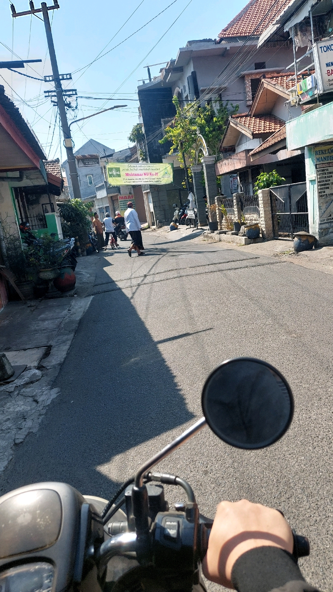 The image shows a street scene in Sidoarjo, Indonesia. A person's hand is visible on the handlebars of a motorcycle, suggesting the image was taken from the perspective of a rider. The street is paved with asphalt and has a shadow cast across it from a building on the left. Several people are walking on the street, some dressed in traditional clothing. Buildings line both sides of the street, with some displaying advertisements and signs. A banner with Indonesian text is visible on the left side of the street, and another sign with the text "MAAF" (meaning "Sorry" or "Excuse me" in Indonesian) is on a building. The sky is clear and blue, indicating a sunny day. The overall impression is a typical street view in a residential or commercial area of a town or city in Indonesia.