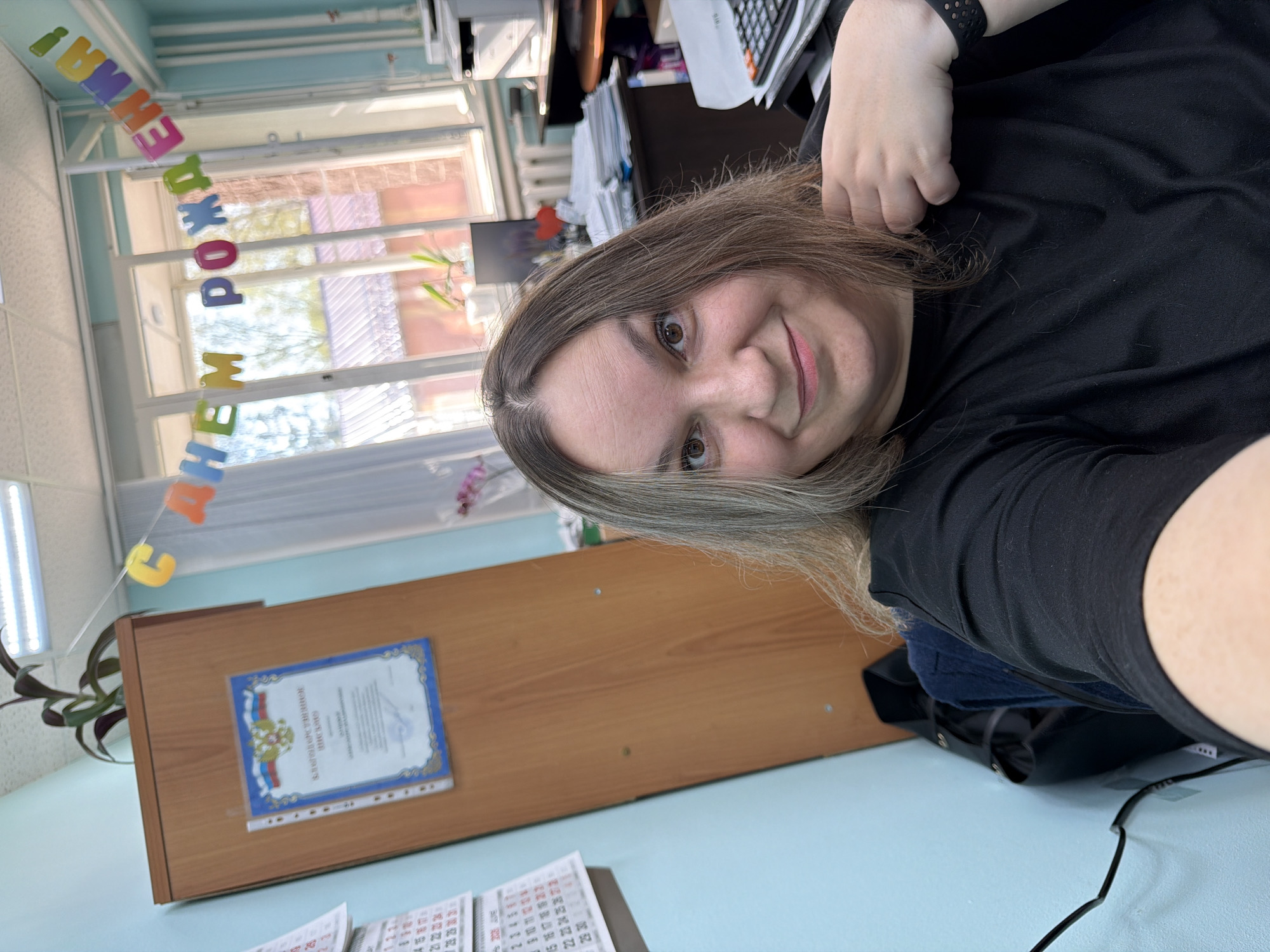 The image is a selfie taken indoors. A woman with light brown hair and a black shirt is facing the camera. Her hand is visible at the top right of the frame, and a black smartwatch is on her wrist. The background shows an office setting. A window with a banner written in Russian hangs above a wooden desk. A certificate in a frame is mounted on the desk. A calendar is visible on the desk. The office seems to be in Chaikovsky, Russia as evidenced by the certificate and language on the banner.