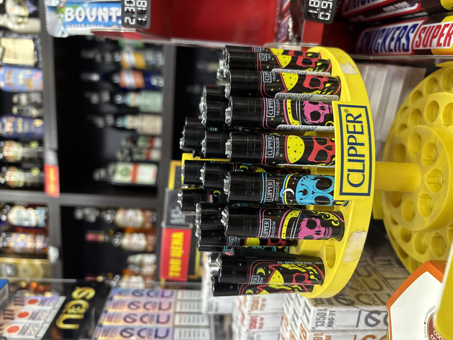 The image depicts an interior view of a retail establishment located in Sofia, Bulgaria. The primary subject is a yellow, two-tiered circular display stand holding multiple black Clipper brand reusable lighters. Each lighter features colorful skull graphics, with some identifiable as "Black Magic Skulls." The top tier of the stand is populated with lighters, while the bottom tier is empty. A "CLIPPER" logo sign is affixed to the front of the display.
To the immediate left of the lighter display, several stacks of white and orange boxes, possibly containing cigarette filters or rolling papers, are visible, marked with numbers like "1350" and "NRG-71." Further left, a blurred black display with a yellow "NBS" logo holds additional boxed products, some labeled "SCU."
Behind the lighter display, a red sign with "ТОП ЦЕНА" (TOP PRICE in Bulgarian) in yellow text is visible. To the right, a red vertical display case features "BOUNTY" and "SNICKERS SUPER" candy bars. Digital price tags on this display show "2.30" and "1.18," indicating prices in Bulgarian Lev. The background consists of dark shelves densely stocked with numerous blurred bottles, consistent with an assortment of alcoholic beverages. No individuals, actions, or direct interactions are visible within the frame. The environment is typical of a convenience store or supermarket aisle.