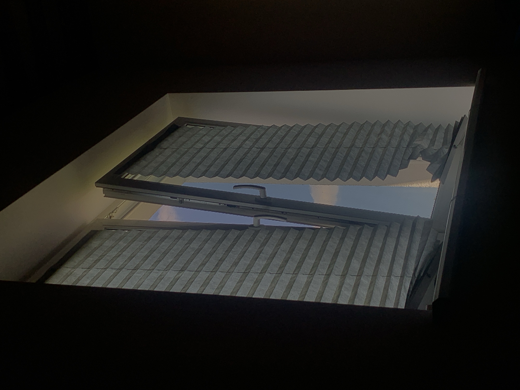 A partially opened window with pleated blinds in a dark room, with a sliver of a cloudy sky visible outside. The window frame is white, and the blinds are a pale gray. One of the blinds appears to be damaged and torn at the bottom. The overall impression is one of a slightly neglected or aged interior. The image does not provide any specific details to pinpoint its location to Minsk, Belarus.
