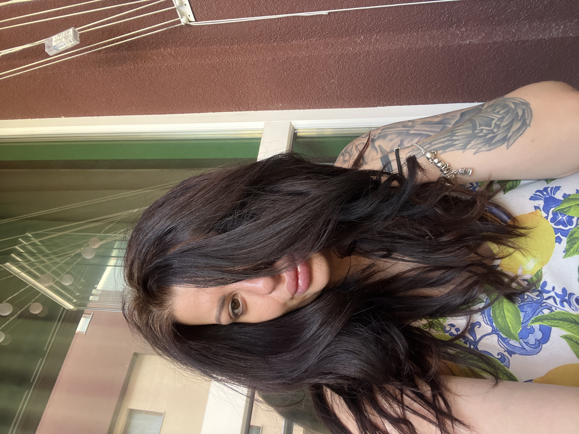 A medium shot captures a woman with dark, voluminous hair framing her face, obscuring the left side of her visage. Her right eye is visible, gazing slightly to the left, while her lips are parted and slightly glossy. She wears a white top adorned with a colorful print of lemons and blue floral patterns. Her left arm, visible on the right side of the frame, features a prominent tattoo. A bracelet with several charms adorns her wrist. The background suggests an outdoor setting, possibly a balcony or patio, with a brown textured wall on the right and a window with a green tint on the left. Reflections in the window show buildings and lights, hinting at an urban environment. The scene is illuminated by natural light, casting soft shadows. There is no indication that the image was taken in Plovdiv, Bulgaria.