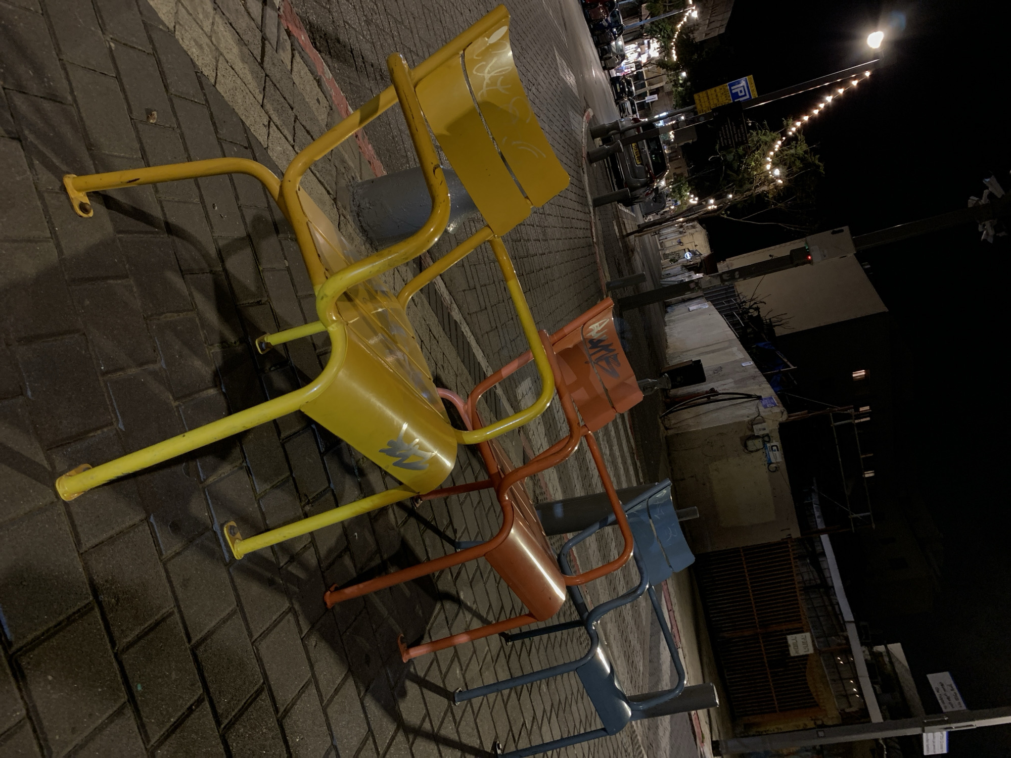 The image depicts a street scene at night, featuring a stack of three metal chairs – yellow, orange, and blue – positioned on a brick-paved sidewalk. The chairs are arranged one behind the other, creating a line that extends into the scene. The background includes a street with vehicles and streetlights, some adorned with decorative lights, suggesting a lively urban environment. The presence of Hebrew signage suggests that the location is in Israel, likely in Tel Aviv given the urban setting and architectural style. Buildings and structures are visible in the background, adding to the depth of the scene. The overall ambiance is that of a quiet night in a vibrant city.