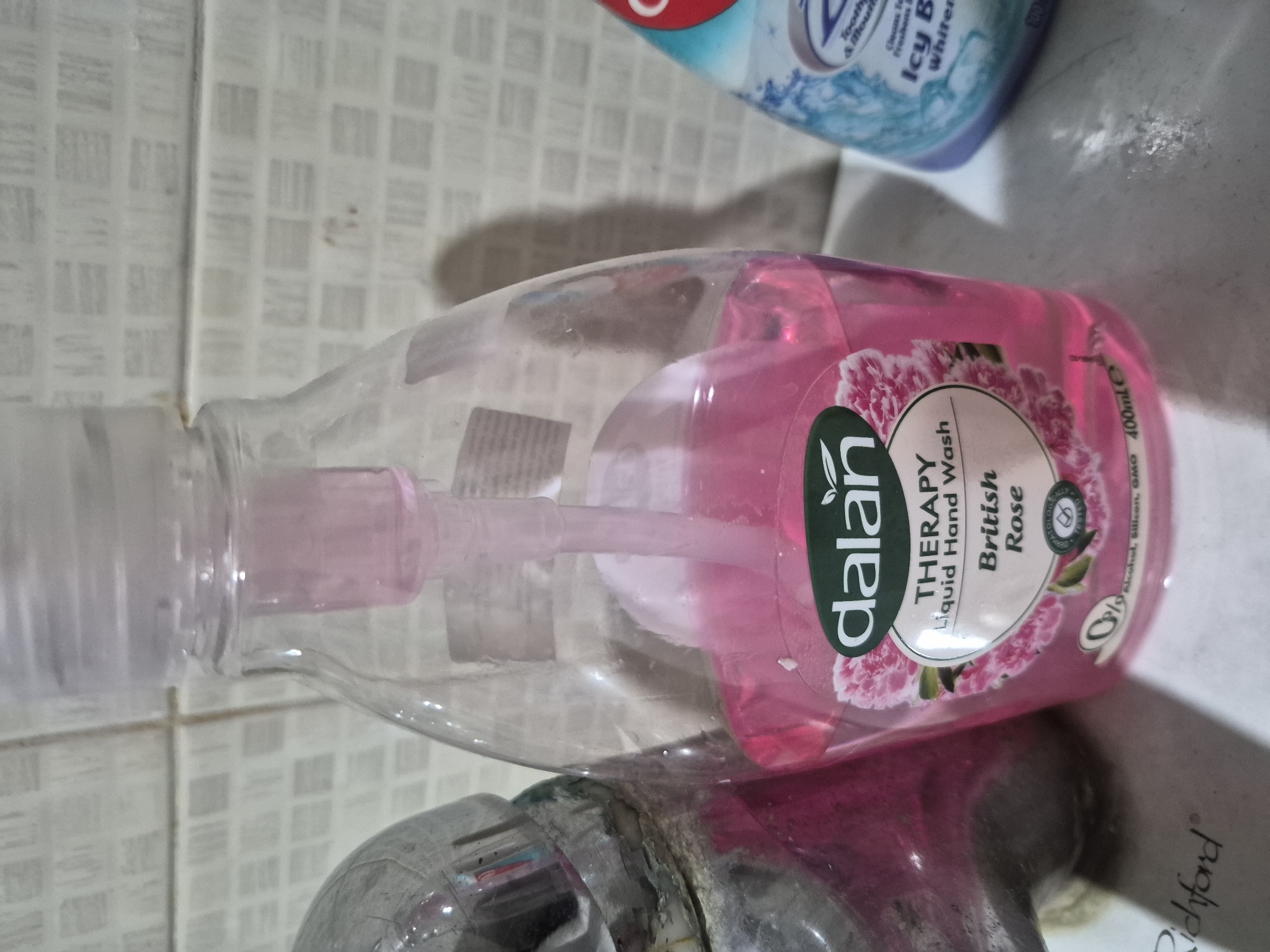 The image displays an indoor scene, likely a bathroom or kitchen sink area, in what appears to be daytime. The main subject is a transparent plastic bottle of "dalan THERAPY Liquid Hand Wash British Rose" containing pink liquid soap, positioned horizontally on a surface next to a metallic faucet. The bottle is about three-quarters full.
Visible text on the hand wash bottle's label includes "dalan", "THERAPY Liquid Hand Wash", "British Rose", "DERMATOLOGICALLY" (partially visible), "0% Alcohol, Silicon, GMO", and "400ml e". The bottle features a pump dispenser.
A metallic faucet, showing some wear and residue, is visible in the lower part of the image. In the upper right corner, a portion of a blue and white bottle is visible, which appears to be a "Colgate" product, possibly mouthwash or toothpaste, with readable text fragments such as "Tooth & Mouth", "Cleans Fresh", "Icy B", and "Whiter".
The background features light-colored tiled walls with a small, grid-like pattern. Below the pink hand wash bottle, the word "Richford" is visible on a white surface, possibly a countertop or a piece of packaging. No people or specific activities are depicted; the scene is a static arrangement of everyday hygiene products. The lighting suggests ambient indoor illumination.