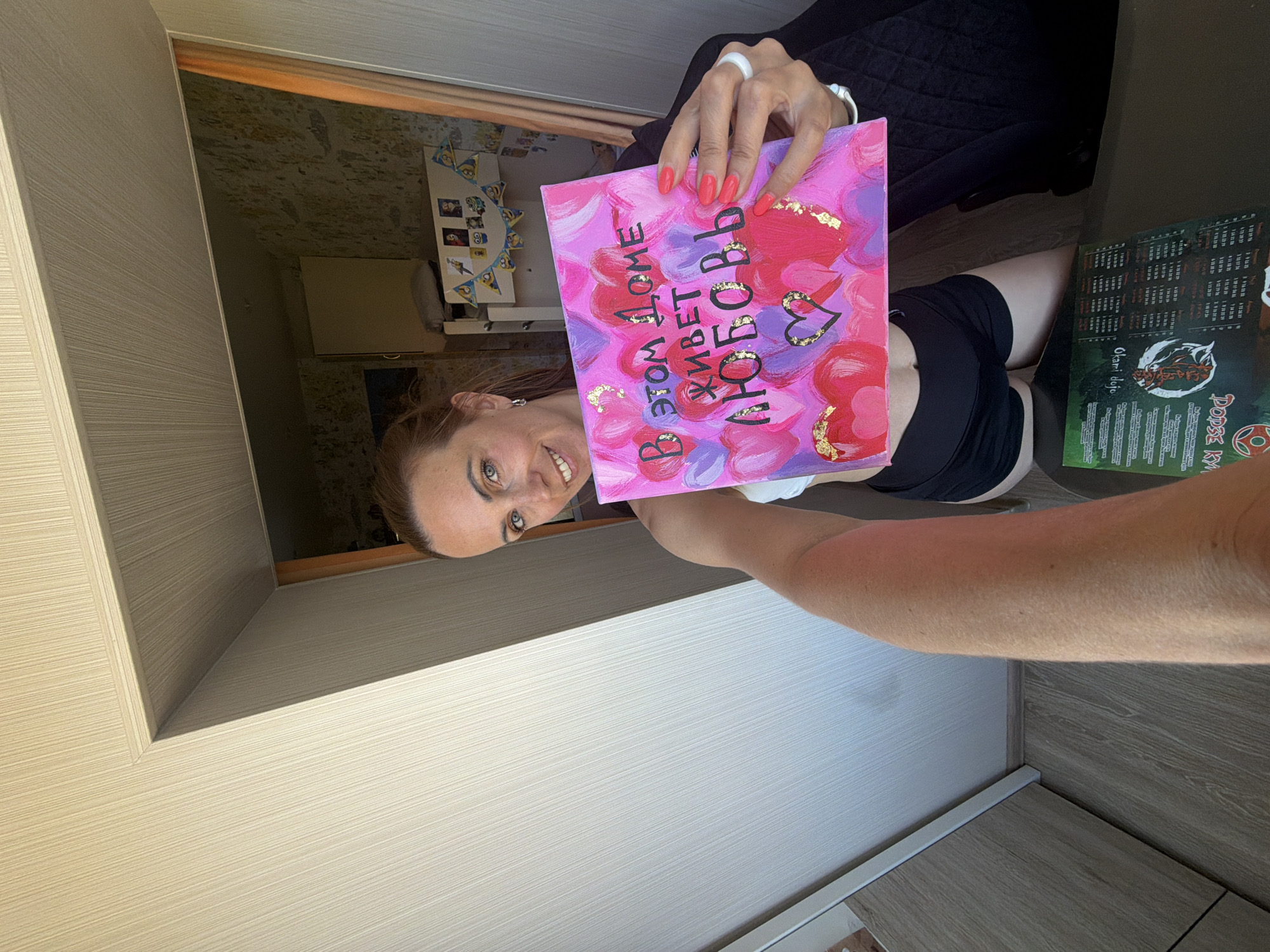 A woman with blonde hair pulled back is holding a pink canvas painted with hearts and the Russian text "В этом доме живет любовь" (Love lives in this house). She is wearing a white sports bra and black shorts. The canvas has gold leaf accents. Behind her, a room is visible with patterned wallpaper, a doorway, and a shelf with decorations that appear to be Minions characters. A menu with Russian text is visible on a dark surface in the foreground, suggesting the location might be a cafe or restaurant. Based on the visual clues and context, the scene appears to be an indoor residential setting, possibly in the city of Birobidzhan, Russia.