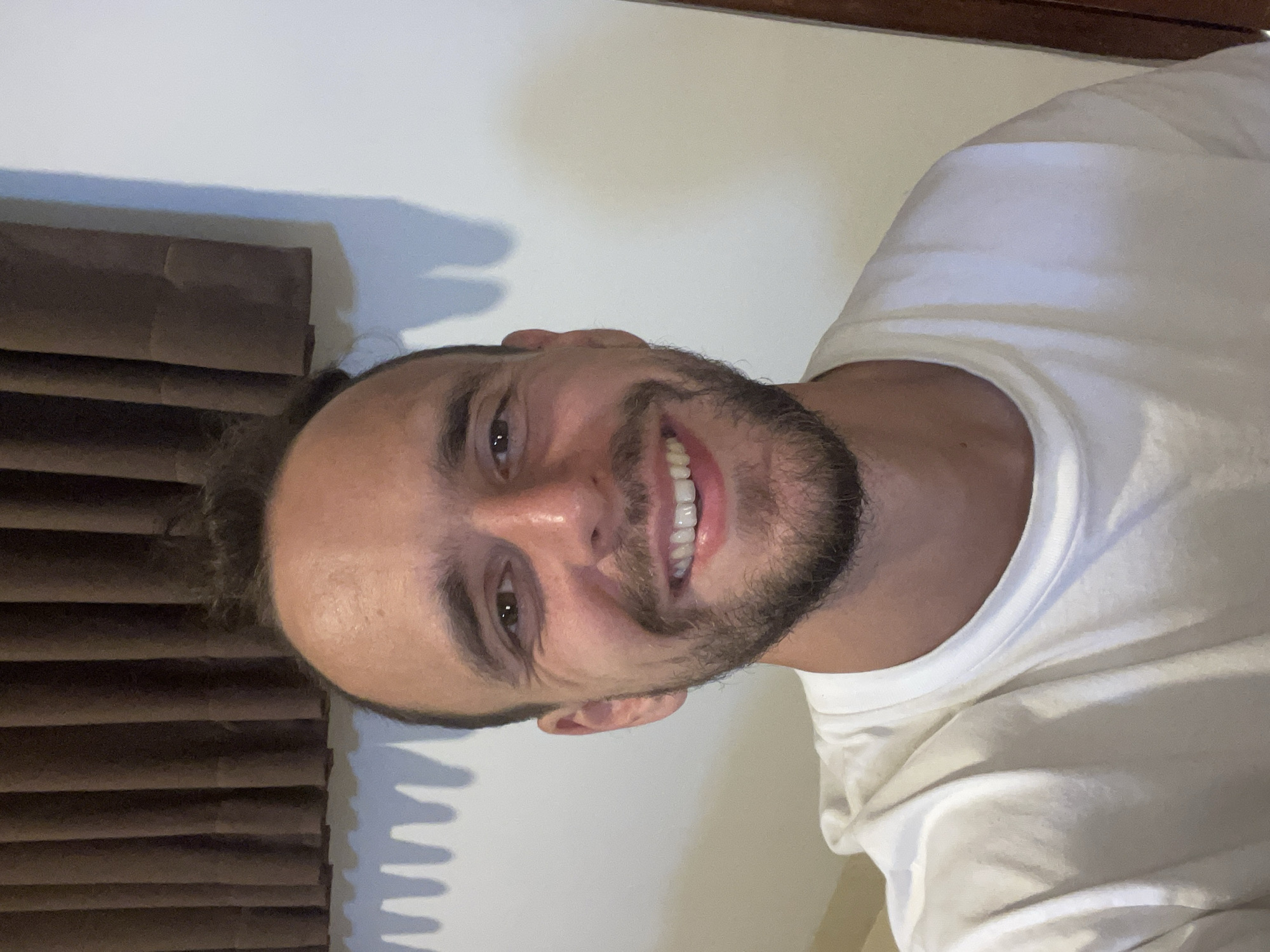 A man with short, dark, messy hair and a beard smiles broadly at the camera. He is wearing a white t-shirt. Behind him, a dark brown curtain is visible, and to his right, a white wall with a shadow of a curtain can be seen. The image appears to be a selfie taken indoors. There is no information in the image to suggest a location in Kediri, Indonesia.
