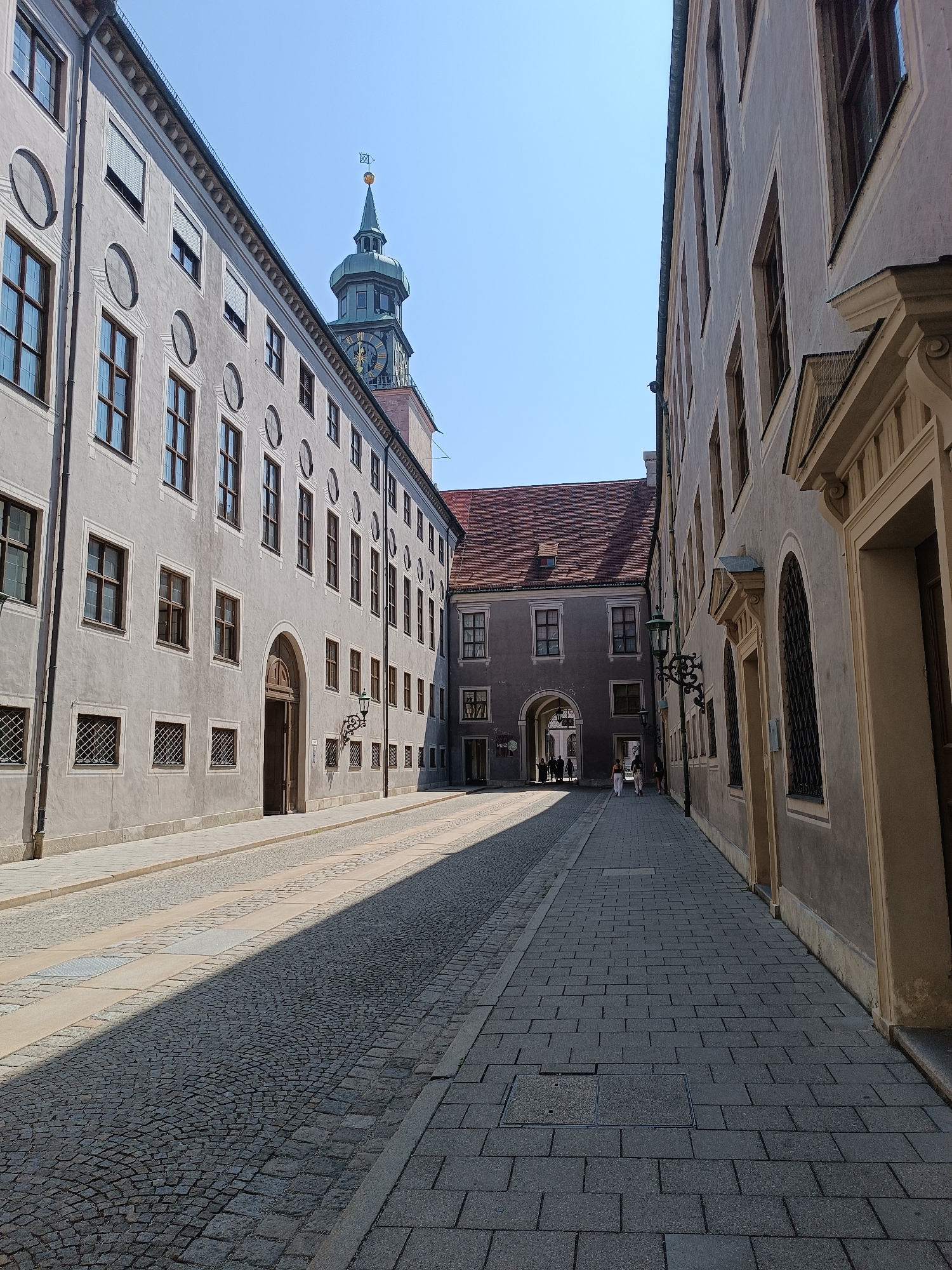 The image shows a view looking down a paved street in Munich, Germany. The street is flanked by buildings on either side, with a tower visible in the distance. The building on the left has a grey facade with numerous windows and decorative circular motifs. The building on the right has a similar facade and architectural style, with arched windows and decorative elements. The street itself is paved with cobblestones, with a sidewalk on the right side. Sunlight casts a distinct shadow across the cobblestone street, indicating it is daytime. A few people are visible walking in the distance, near an arched entryway between buildings. The sky is clear and blue. The overall impression is of a historic European city street.