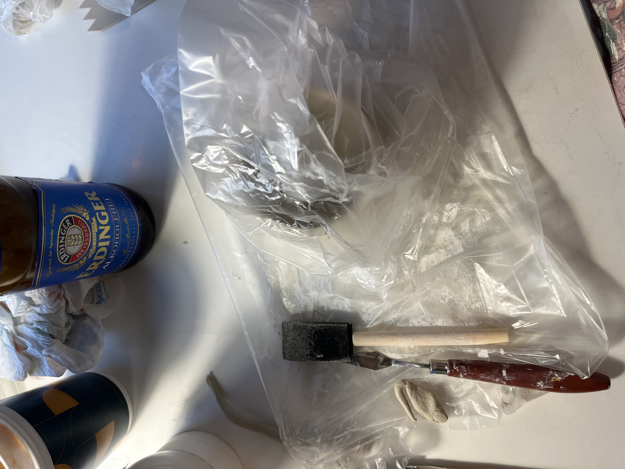 The image displays a collection of items on a white surface, likely a table or desk. In the upper left is a white and blue patterned cup, next to a crumpled white paper towel. To the right of the cup is a brown glass bottle with a blue and yellow label that reads "ERDINGER ALKOHOLFREI."
In the foreground, a plastic bag is spread out, partially covering a beige object, possibly clay or a similar material. Inside the plastic bag, a small, light-colored object resembling a piece of clay is visible on the left side. Next to it are two tools: a sponge brush with a black sponge head and a wooden handle, and a small spatula with a red handle and a metal blade. The spatula has some white residue on its handle.
The lighting suggests an indoor setting with a light source casting shadows. The overall scene appears to be a workspace or a preparation area for some craft or artistic activity. There are no people or animals visible in the image, and no discernible landmarks that would indicate a specific location in Limassol, Cyprus.