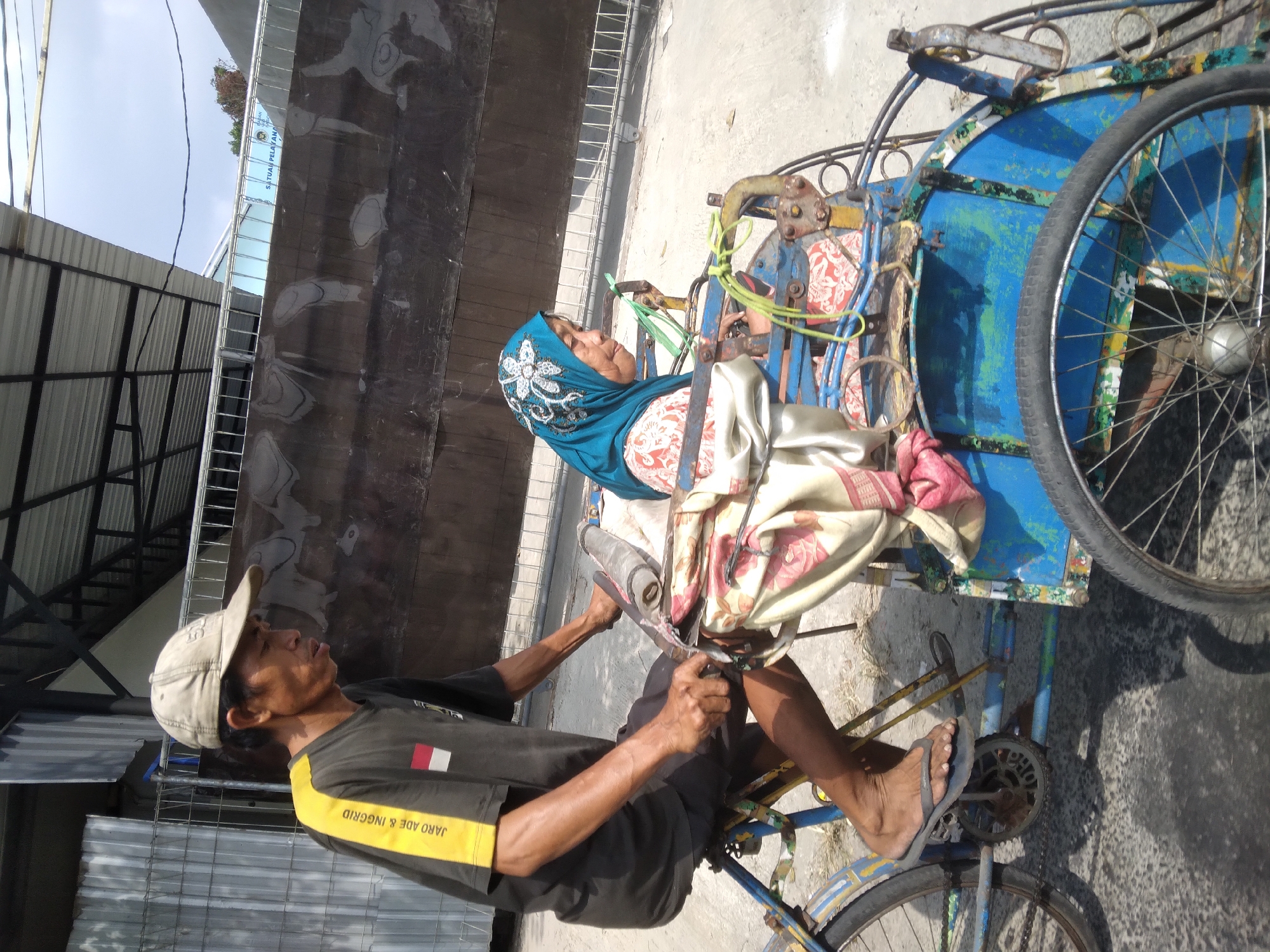 In Purwokerto, Indonesia, a man with a tan baseball cap is pedaling a pedicab. He is wearing a dark t-shirt with a yellow stripe on the left sleeve and the Indonesian flag on the chest. A woman wearing a blue headscarf and a floral patterned shirt is seated in the pedicab behind him. The pedicab is blue and yellow and appears to be in use. The background shows a building with a corrugated metal roof and a gray wall with a sign that reads "SATUAN PELAYANAN." The scene suggests a typical day in a bustling Indonesian city.