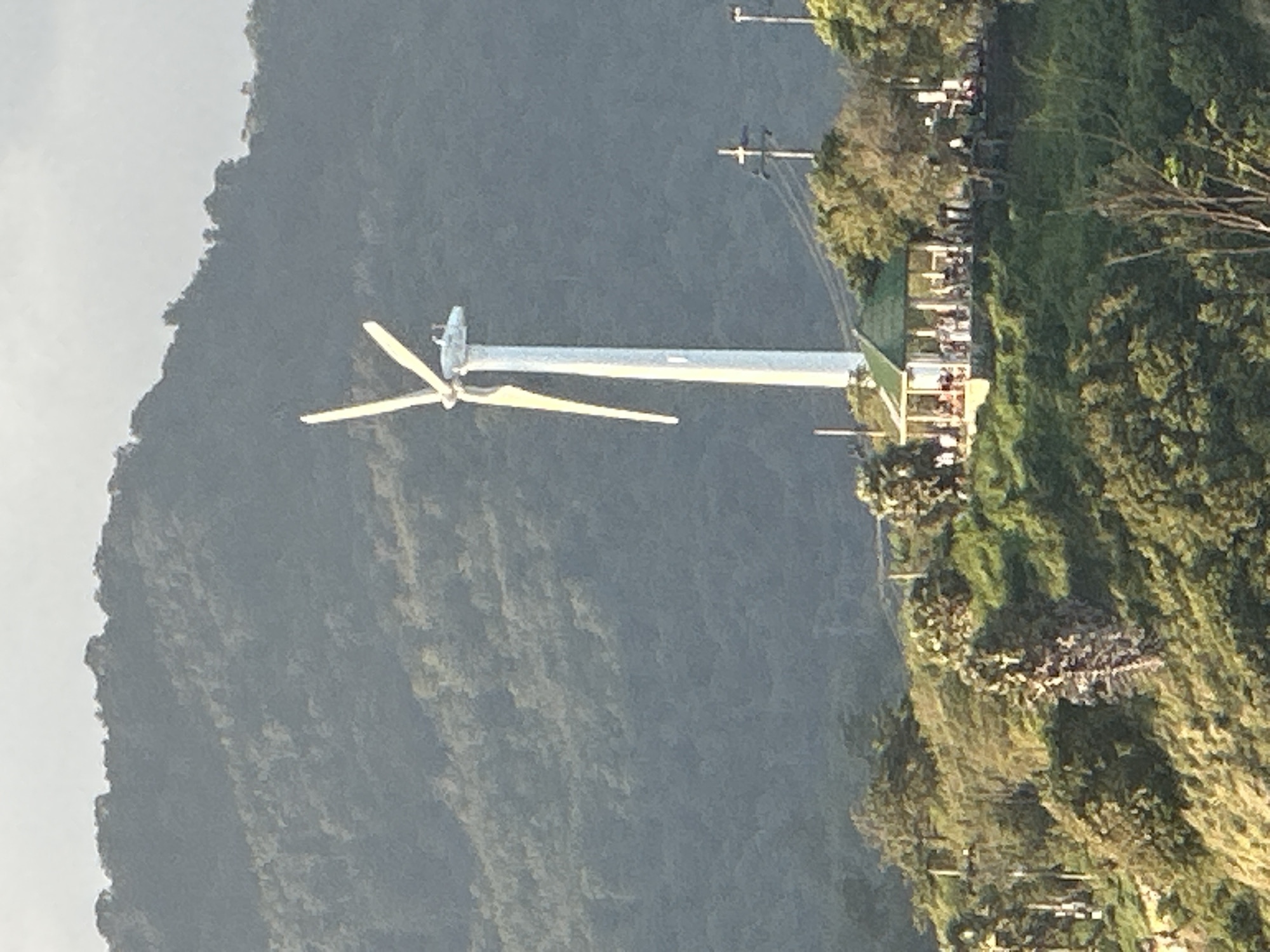 In Rawai, Thailand, a prominent white wind turbine stands tall on a green, treed hillside. Its blades are angled upwards and slightly to the left, positioned against a hazy, light-colored sky. At the base of the turbine, a gazebo with a green roof is visible, where numerous people are gathered. The gazebo is surrounded by lush vegetation and trees. In the background, a densely forested mountain slopes upwards. Electrical poles and wires are also present in the mid-ground, near the gazebo. The scene is illuminated by natural light, suggesting it is daytime.