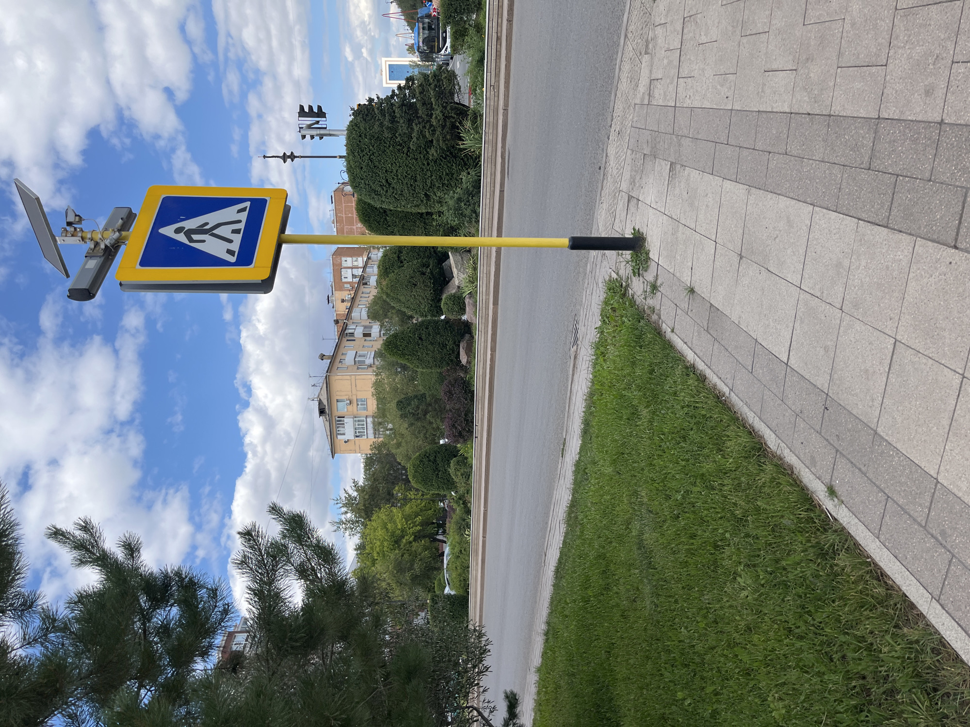 A pedestrian crossing sign is visible on a yellow pole on a sidewalk in Astana, Kazakhstan. The sign is a blue square with a yellow border and a white silhouette of a person walking. Above the sign is a solar panel and what appears to be a camera. The sidewalk is made of grey paving stones. To the left of the sidewalk is a patch of green grass and a portion of a pine tree. Beyond the sidewalk is a road and then a landscaped area with bushes and trees. In the background, there are buildings, a traffic light, and a bus. The sky is blue with white clouds.