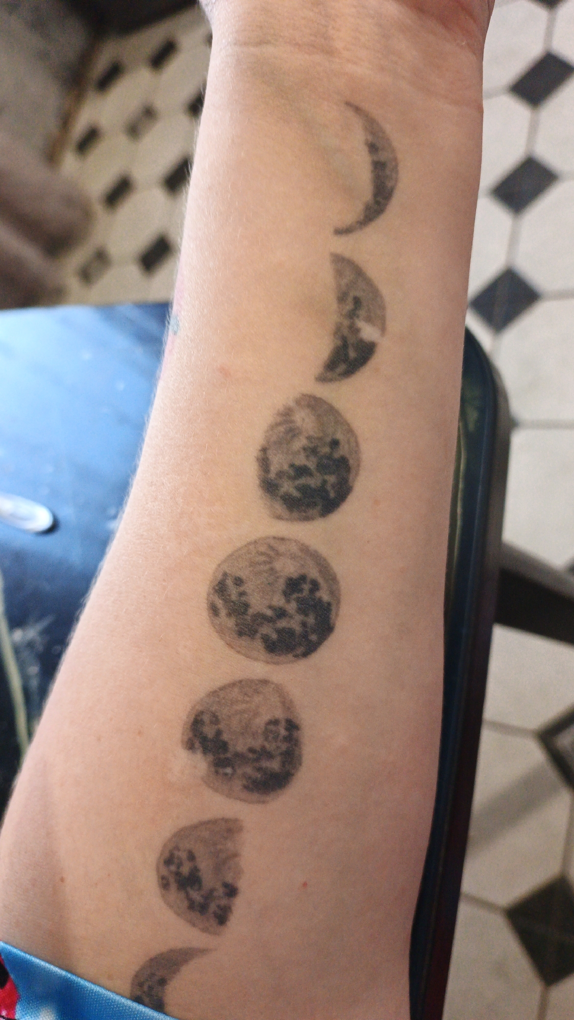 The image shows a forearm with a tattoo of moon phases. The tattoo starts with a crescent moon near the wrist and progresses to a full circle, then back to a crescent, indicating a cycle. The skin is fair, and the tattoo ink appears to be black. The forearm is positioned vertically, with the wrist at the bottom of the frame. In the background, a tiled floor with a black and white diamond pattern is visible, along with a blurred glimpse of a blue surface, possibly a table. The lighting suggests an indoor environment. The location of Drezna, Russia, is not discernible from the image itself and would require external information to ascertain.