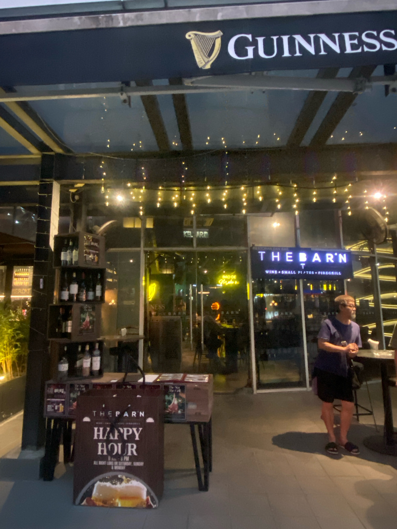 The image shows the exterior of a bar called "THE BARN" in Kuala Lumpur, Malaysia, at night. A Guinness sign is visible above the entrance, and string lights are hung across the facade. In the foreground, a sign advertising "HAPPY HOUR" is displayed. A man is standing near a table outside the bar. The bar appears to have a modern, open-air design with glass windows.
