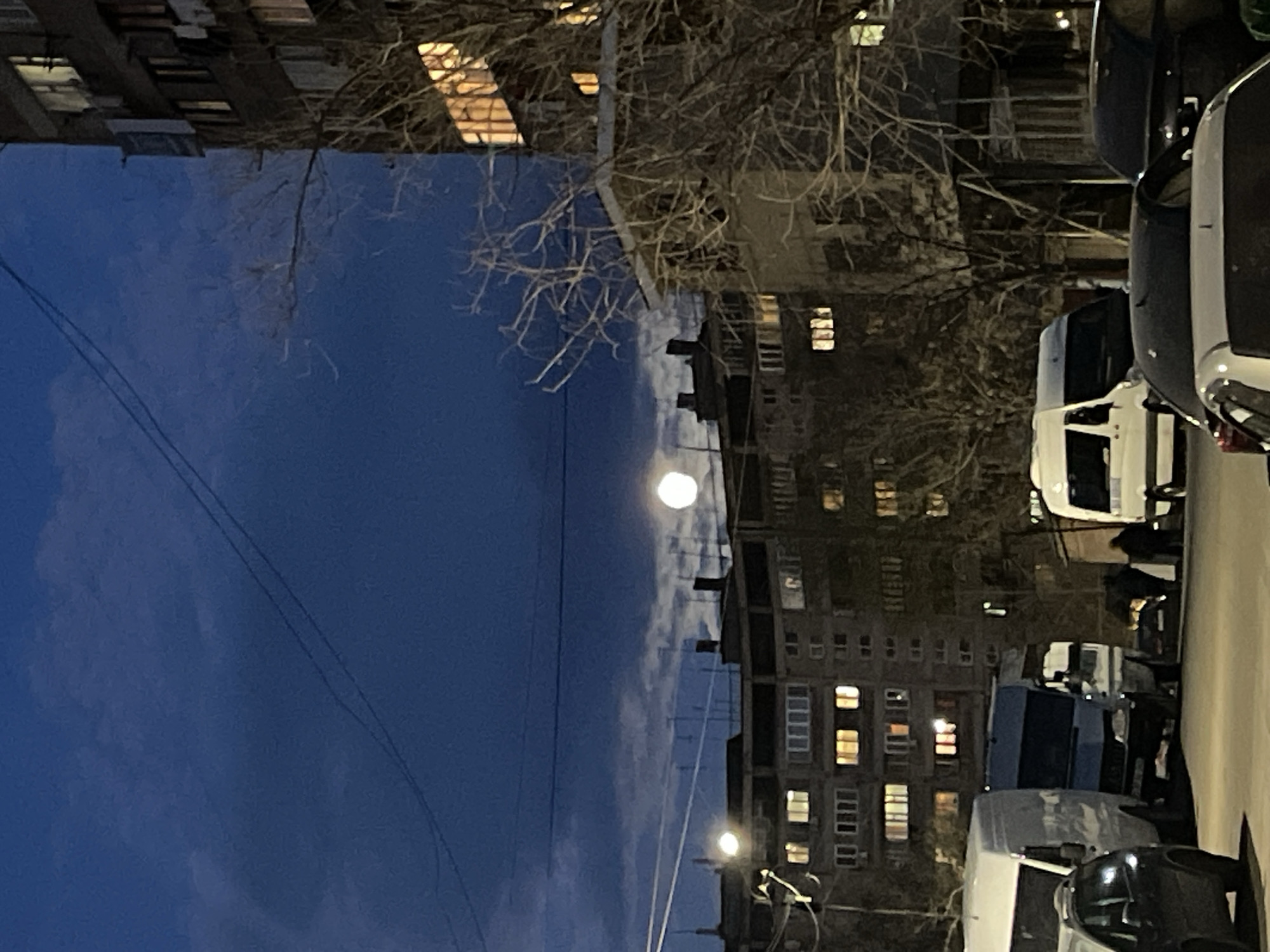 The image shows a Yerevan street scene at night. Apartment buildings with lit windows line the street. In the sky is the moon. Bare tree branches are visible. Several cars and vans are parked along the right side of the street, some with headlights on. At least one person is walking by the vehicles. Overhead power lines are visible in the dark blue sky.