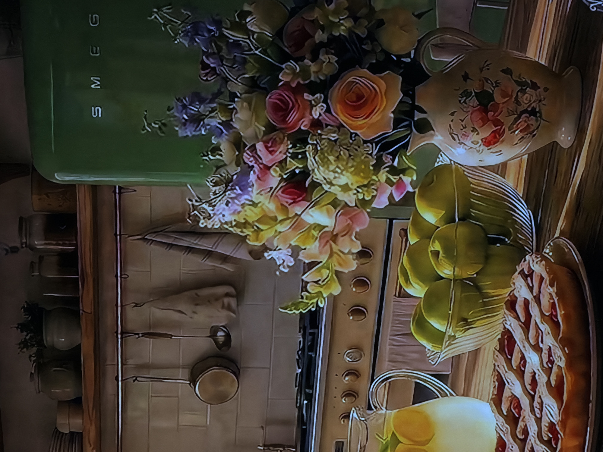 This image captures a vibrant and artfully arranged kitchen scene, likely staged for aesthetic appeal. On the left, a retro-style, pale green SMEG refrigerator stands prominently, adorned with a large, colorful bouquet of flowers featuring roses, delphiniums, and other blossoms in shades of pink, orange, yellow, and purple. To the right of the flowers, a cream-colored pitcher with delicate floral paintings adds to the charm.
Below this arrangement, a clear bowl overflows with bright green apples, positioned next to a rustic-looking baked tart or pie with a lattice crust and a red fruit filling. In the bottom right corner, a clear glass pitcher holds what appears to be lemonade or infused water with lemon slices.
The background reveals a vintage-style gas range with multiple burners and control knobs. A light-colored tiled wall serves as a backsplash, and a wooden shelf displays several jars or pots. Various cooking utensils, including a ladle and a small pot, hang from hooks on the wall, suggesting a functional yet decorative kitchen space. The overall impression is one of homeliness, freshness, and rustic elegance. No people are visible, and no specific activity is taking place, presenting a static, inviting domestic scene.