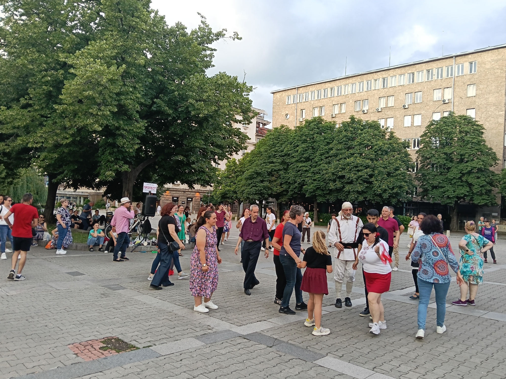 This image depicts a daytime public gathering in Battenberg Square (Площад Батенберг) in Sofia, Bulgaria. Approximately thirty-five individuals are visible, primarily adults of various ages, with a few children present. The scene focuses on a group of people engaged in what appears to be a traditional Bulgarian folk dance (horo) on a paved plaza.
In the foreground and midground, multiple individuals are standing in a loose circular or linear formation, some holding hands, indicating dance movements. One man is dressed in traditional white Bulgarian attire with a dark belt and white cap. Another woman near him holds a small red and white flag. Other participants wear casual clothing, including t-shirts, dresses, and pants. Some individuals observe the activity, with a few seated on a low wall or grass on the left side of the frame.
To the right, near the dancing group, a white sign with the Cyrillic word "ПРОТЕСТ" (PROTEST) in red letters is mounted on a stand above a black speaker system. A bicycle is leaning against the speaker stand.
The environment consists of a light-colored paved square with distinct square and rectangular concrete tiles. Large, mature deciduous trees with dense green foliage are prominent throughout the scene, providing shade. In the background, a multi-story building, characteristic of the former Party House now part of the National Assembly complex, is visible. This building has numerous rectangular windows and multiple exterior air conditioning units. The sky is overcast with a uniform grey cloud cover. The overall impression is an organized public event combining cultural activity with potential elements of public expression, indicated by the "PROTEST" sign.