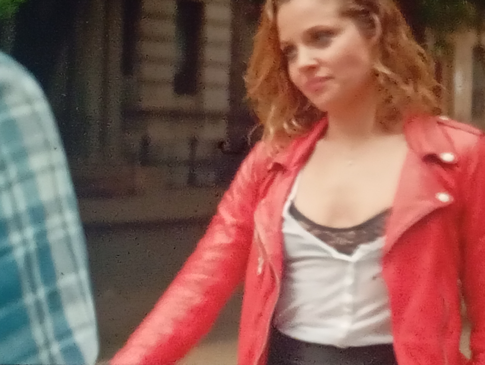 In a medium shot, a woman with curly blonde hair is seen from the chest up, facing left. She wears a red leather jacket over a white blouse with black lace trim. Her head is turned, and she looks towards the left side of the frame. To her left, partially visible, is another person wearing a blue and white plaid shirt. The background appears to be an outdoor setting with blurred foliage and a building structure, potentially in Sofiivska Borshchahivka, Ukraine. The image is grainy and slightly out of focus.