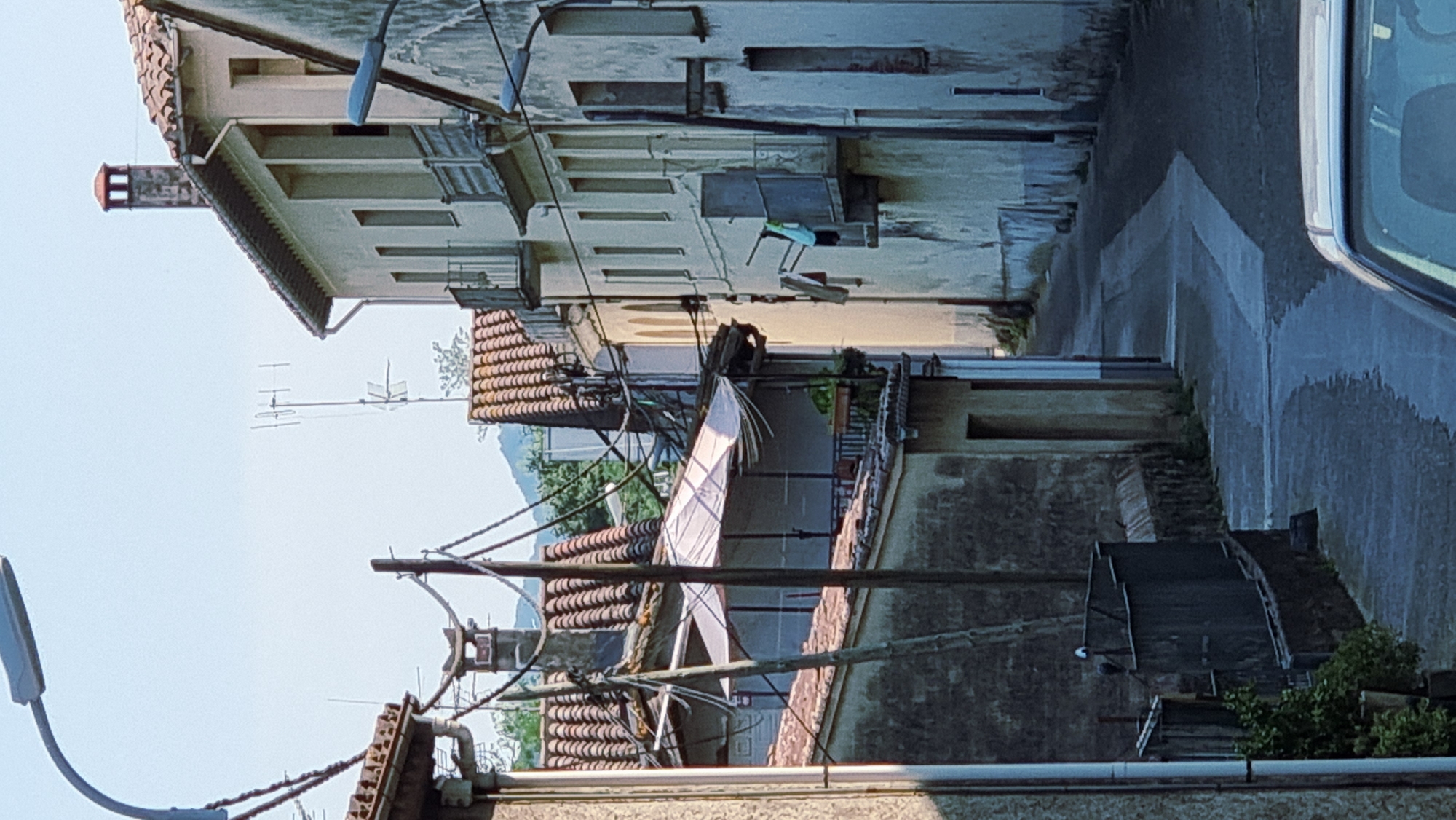 The image shows a narrow street in the town of La Canya, Spain, flanked by buildings with tiled roofs. A streetlamp is visible in the upper left corner. Telephone poles and wires crisscross the scene, with antennas also present on some rooftops. The buildings are primarily light-colored with balconies and windows. Some laundry is hanging from a balcony on the right side. The street itself is paved and appears wet in places, suggesting recent rain or washing. Vegetation is present on the left side of the street, including potted plants and bushes. The scene is captured during daylight with a clear sky.