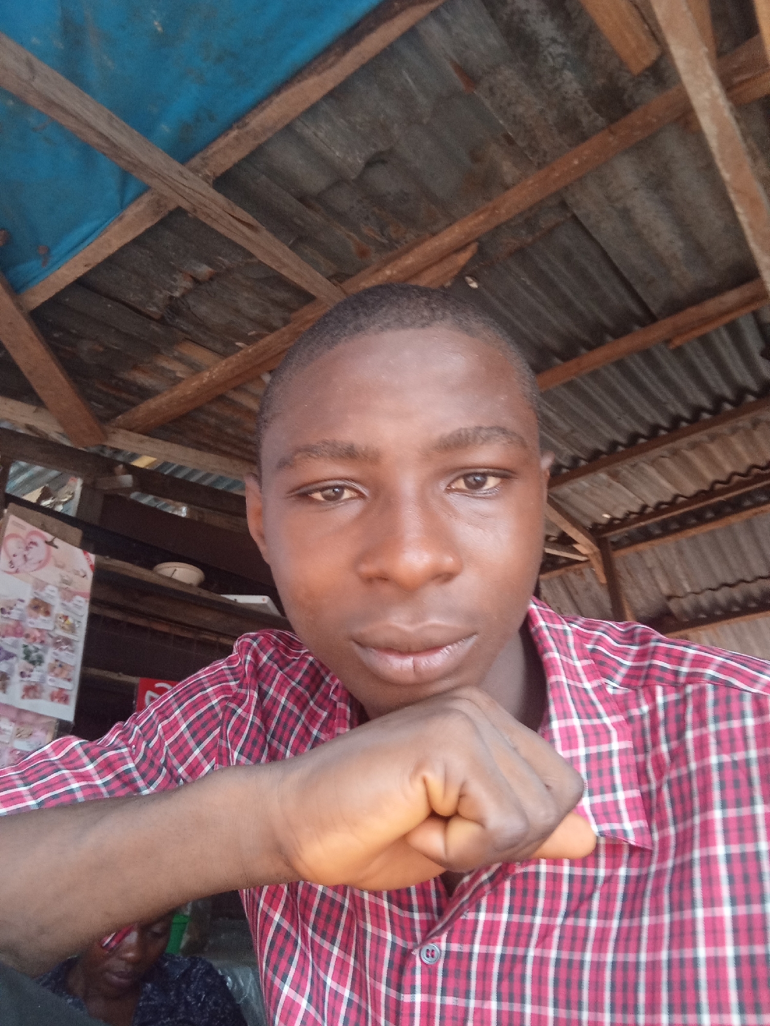 The media file is a close-up, eye-level selfie of a young Black person wearing a red and white checkered shirt. They are resting their chin on their fist, looking directly at the camera with a neutral expression. The background suggests an outdoor or semi-outdoor setting, possibly a market or open-air structure, with a corrugated metal roof and wooden beams visible above. A blue tarp is also present above the beams. To the left of the subject, there is a wall with several framed items or posters, some of which appear to have text and images. Below and slightly to the left of the subject's face, another person's head and shoulder are visible, appearing to be wearing a dark, textured garment. The lighting suggests it is daytime.