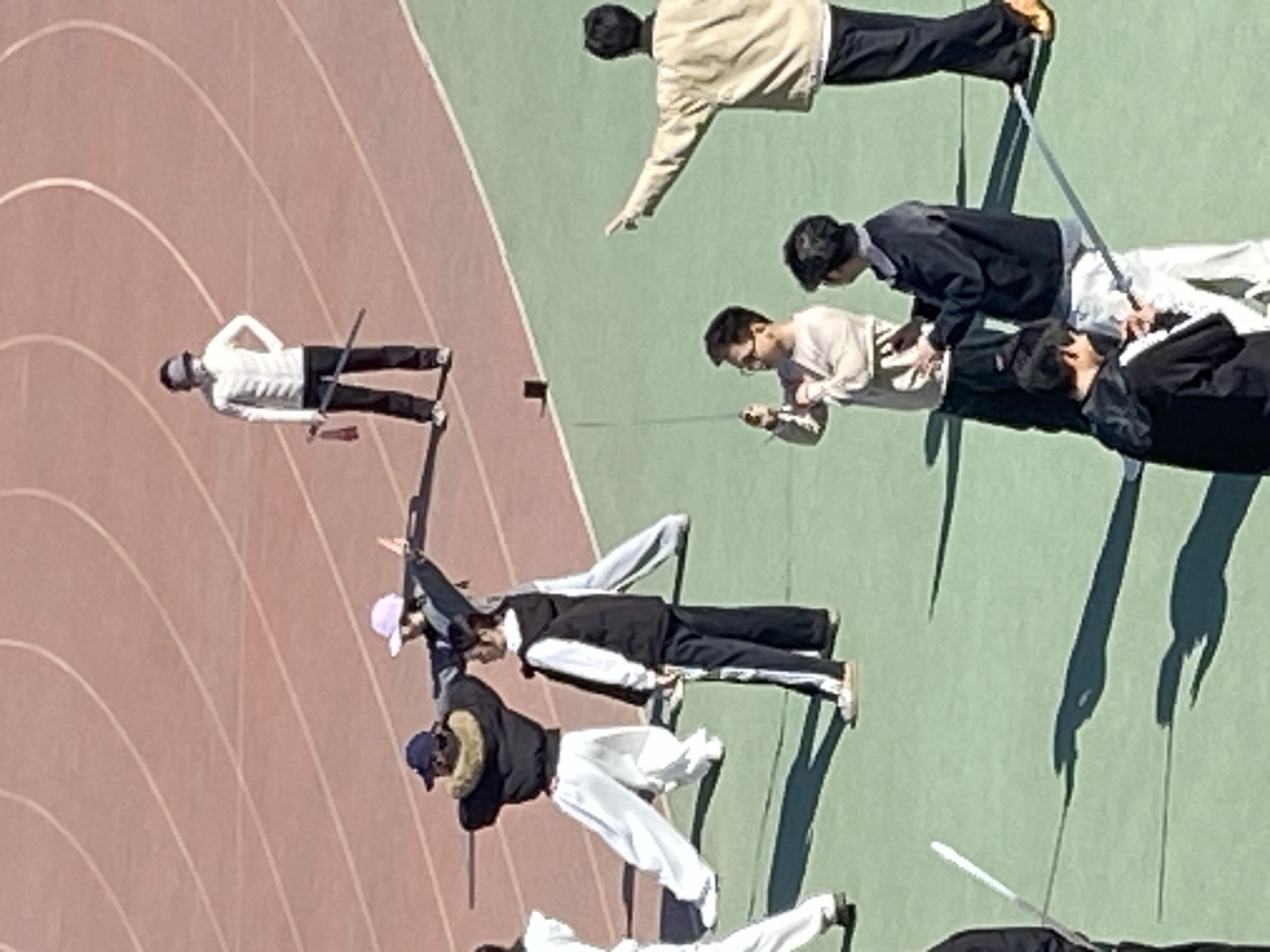 The image shows a group of people on a track and field area, possibly in Zhongtun, China, based on context not contained in the image. The track is visible with multiple lanes marked in a reddish-brown color, transitioning to a green area. Multiple individuals are positioned on the track, each holding what appears to be a fencing foil or similar thin rod. The people are wearing a variety of clothing, including jackets, pants, and hats. They seem to be engaging in some sort of exercise or training activity involving the rods. The scene is captured from an elevated viewpoint, looking down at the activity.