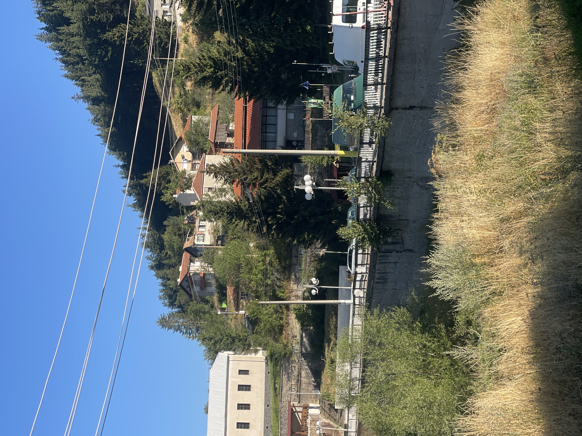 The image shows a hillside village in Manastir, Bulgaria. The village is situated on a steep slope, with houses and trees dotting the landscape. A road runs along the top of the hill, with several vehicles visible. The sky is clear and blue, and the sun is shining brightly. There is a river or ravine in the foreground, filled with dry grass.