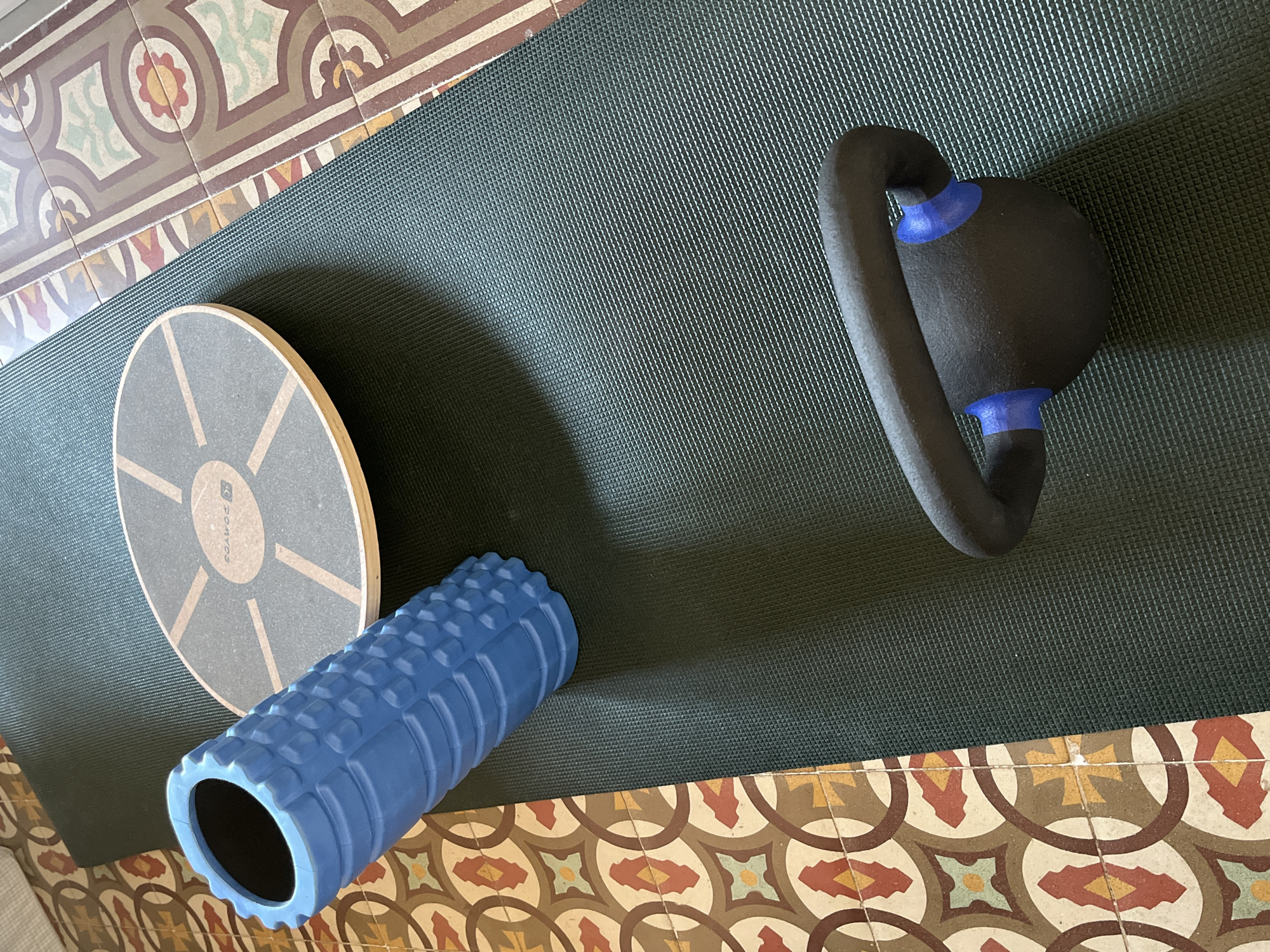 This image captures a top-down view of exercise equipment laid out on a dark green yoga mat, which rests on a distinctly patterned tiled floor. The mat occupies the central and right portions of the frame, while the vibrant floor tiles border it along the left and bottom.
The primary subjects on the mat include a round wooden balance board, a blue textured foam roller, and a black kettlebell with blue accents. The balance board, positioned on the left side of the mat, features a light gray top surface with radial beige lines and the visible brand name "DOMYOS." Next to it is a hollow, bright blue foam roller with a prominent textured grid pattern. Towards the upper right, a black kettlebell is visible, distinguished by its ergonomic shape and royal blue connecting pieces where the handle meets the weight.
The floor beneath the mat is covered with ornate, traditional tiles, showcasing intricate geometric and stylized floral motifs. The color palette of the tiles includes shades of cream, terracotta, light green, yellow, and various browns, characteristic of Catalan modernism or similar architectural styles often found in Barcelona, Spain. The scene is brightly and evenly lit, suggesting daytime. No people are present, but the arrangement of the fitness equipment implies a setting for a home workout, balance training, or physical therapy session.