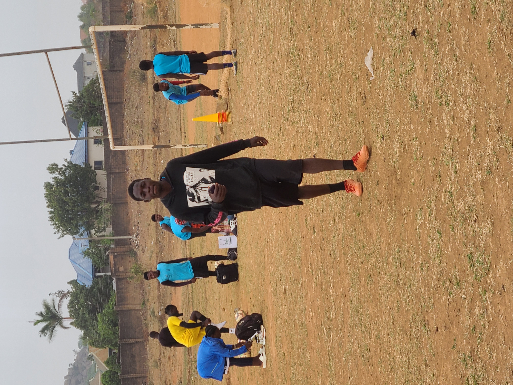 The image depicts a group of people on a grassy field in Barikin Jirga Summa, Nigeria. A soccer goal is visible in the background. A man in a black sweatshirt and orange shoes stands in the foreground. Other people are scattered around the field, some stretching or sitting, wearing blue, yellow, and black clothing. Bags and a cone are also present. The background includes buildings, trees, and a clear sky.