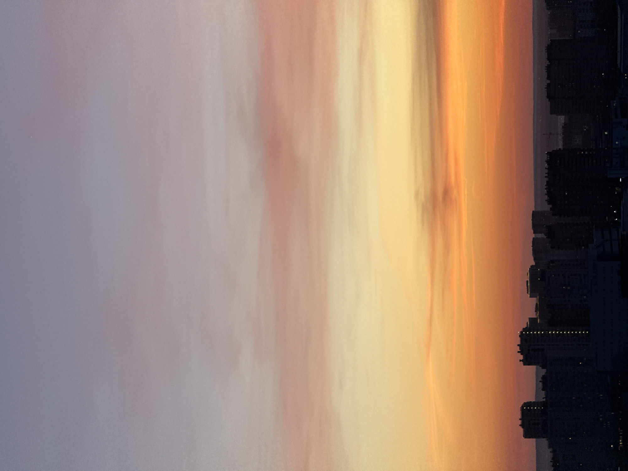 The image depicts a sunset over what appears to be the city of Perm, Russia. Silhouetted buildings are visible along the horizon, suggesting an urban environment. The sky transitions from shades of purple and pink on the left to orange and yellow on the right, indicating the setting sun. The perspective suggests the photograph was taken at an elevated position, possibly from a high-rise building or hill overlooking the city.