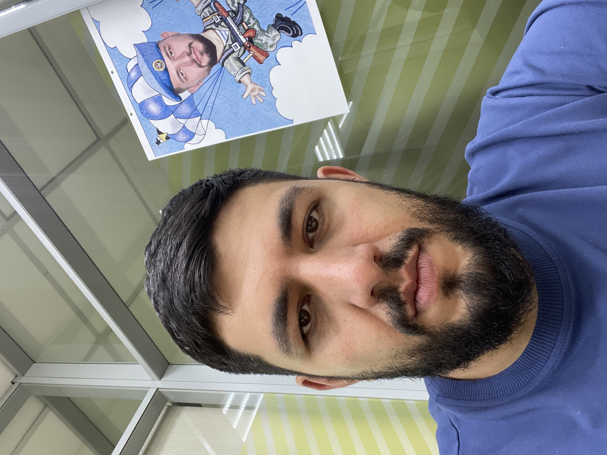 A close-up, indoor selfie features a man with dark hair and a beard, wearing a blue sweater. Above and slightly behind him is a cartoon drawing pinned to the wall. The cartoon depicts a man resembling the selfie subject, dressed in military fatigues and wearing a blue paratrooper beret, descending with a parachute. The wall behind the man has alternating vertical stripes of green and white. Behind and to the left of the man and the drawing is a grid of square windows or partitions. The location is most likely an office space in an urban environment. The city of Krasnoyarsk, Russia is not discernible from the image, so that cannot be determined.