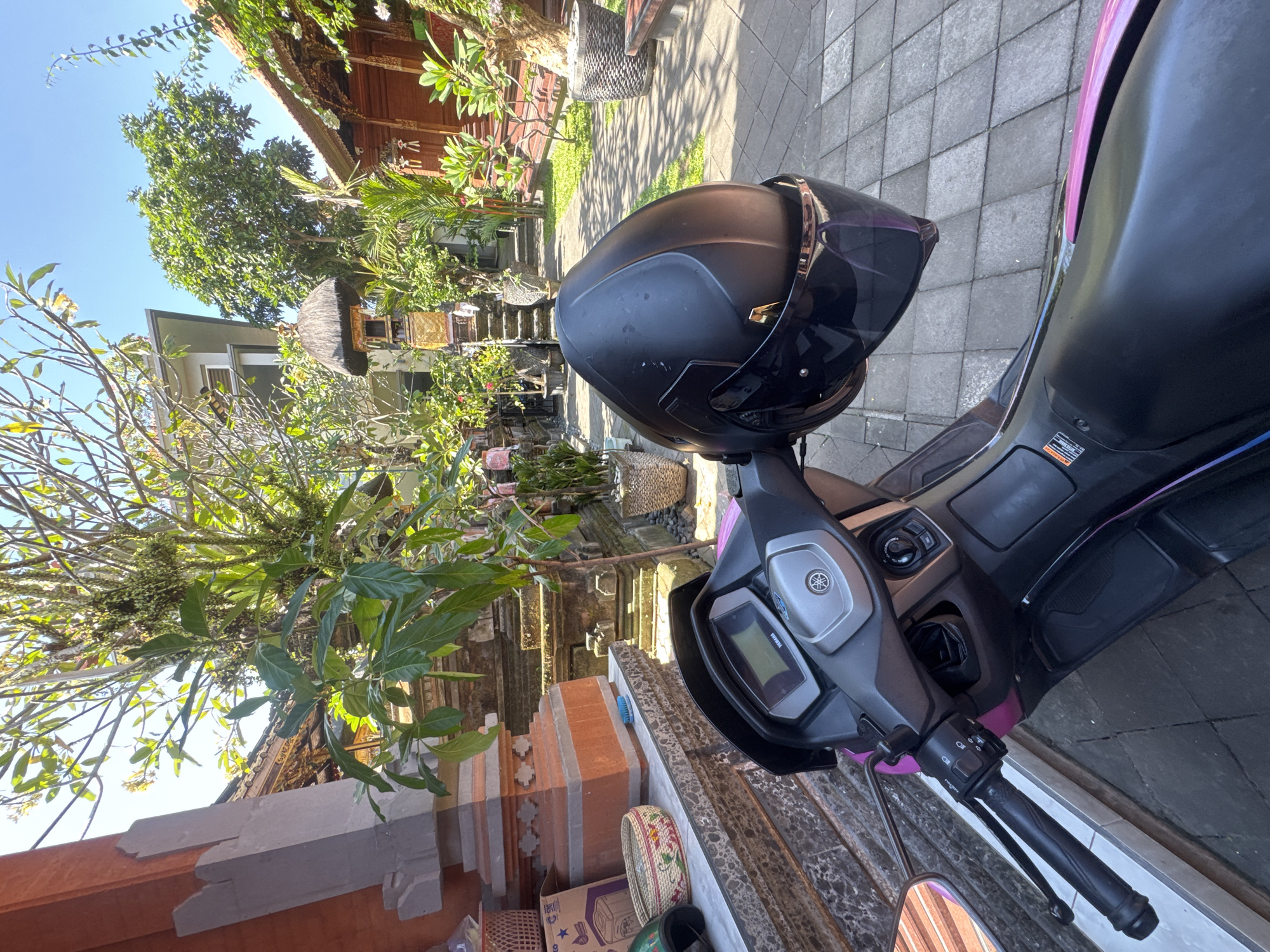 A black motorcycle helmet rests on the handlebars of a black scooter, parked on a paved area. The scooter's dashboard, featuring a digital display and a Yamaha logo, is visible. The scene is set in what appears to be a residential or temple courtyard in North Kuta, Indonesia, characterized by traditional Balinese architecture with intricate carvings and wooden structures, adorned with lush green vegetation. The sunlight suggests it is daytime, likely morning or afternoon, with clear skies. In the background, there are stairs leading up to a building, and several potted plants and decorative items are scattered around. The overall impression is one of a serene, possibly tropical, environment. No people are visible in the image.