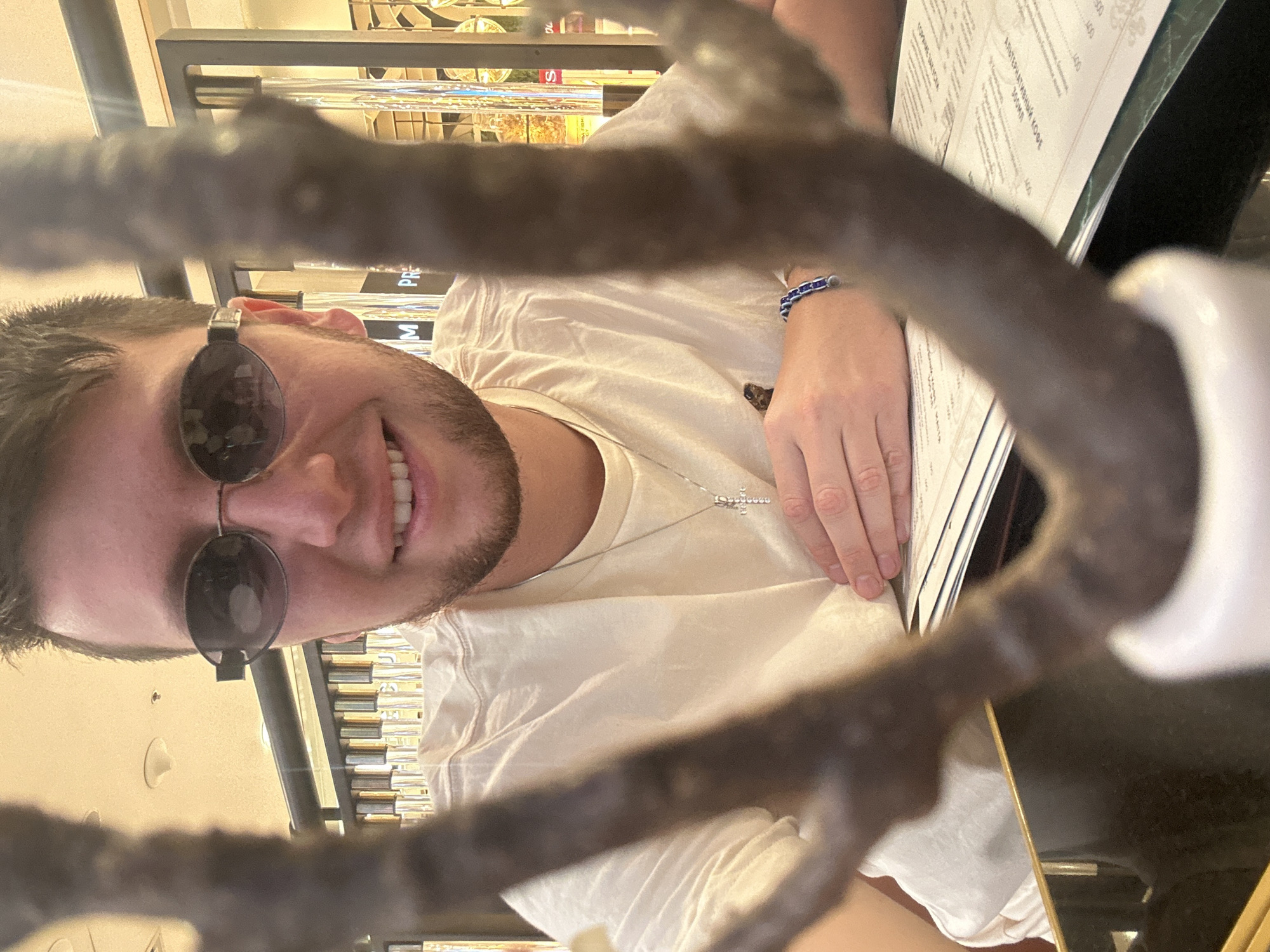 A young man is pictured from the chest up, leaning forward and smiling at the camera through a dark, blurred branch. He wears a light-colored t-shirt and an oval-shaped pair of dark sunglasses. A silver cross necklace with small stones is visible on his chest, and a blue beaded bracelet is on his left wrist. He is sitting at a table with a menu in front of him. The background appears to be an indoor setting, possibly a restaurant or bar, with shelves containing various items, some of which are illuminated. The lighting is warm, suggesting artificial light.