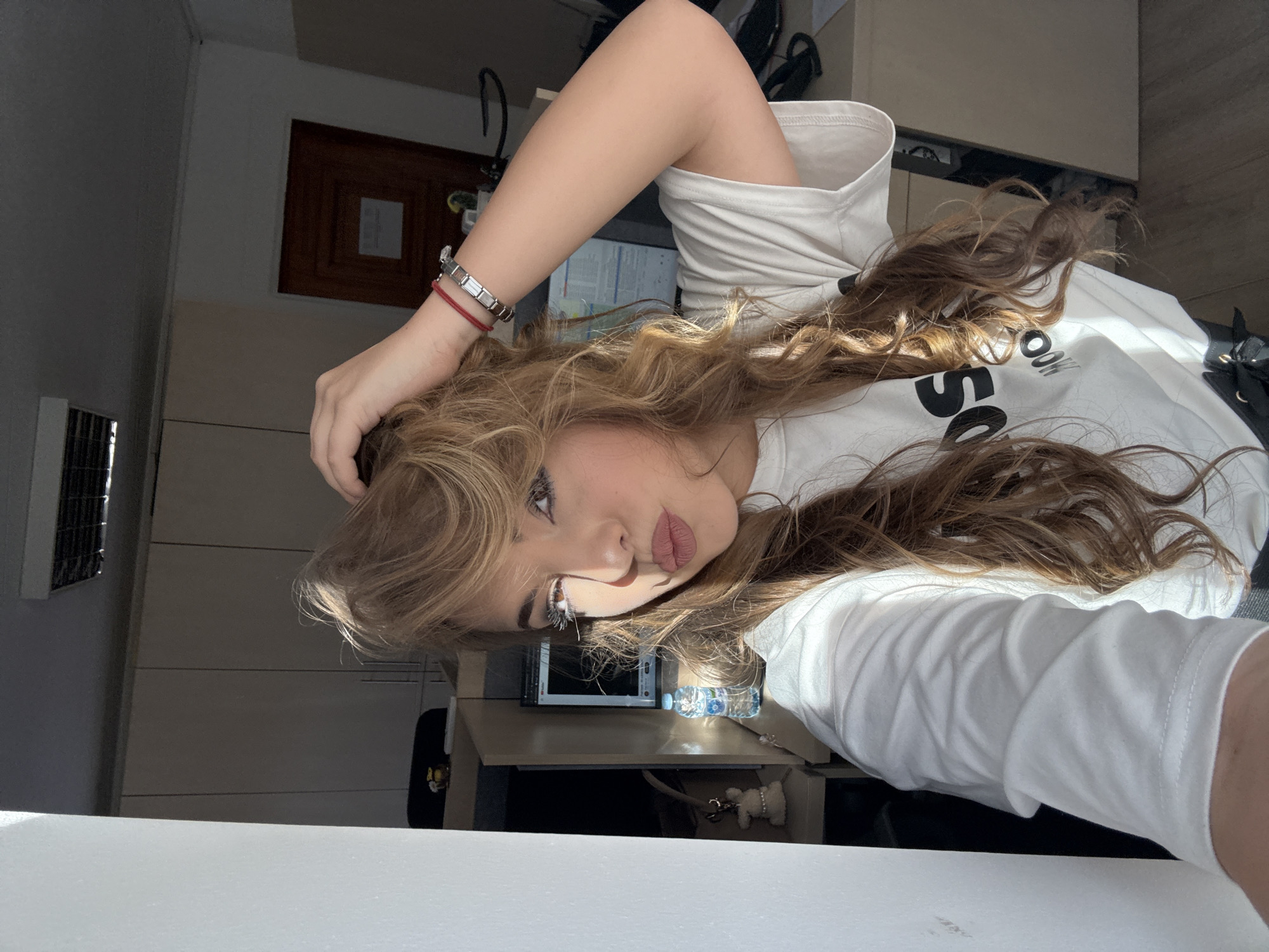 A young woman lies on her side in a room illuminated by sunlight. She wears a white shirt and bracelets on her left wrist. Her long brown hair is spread around her. Behind her is a desk with a computer monitor, a microwave, and a water bottle. The background shows a door and a vent. The location is potentially in Sofia, Bulgaria, but this cannot be confirmed solely from the image.