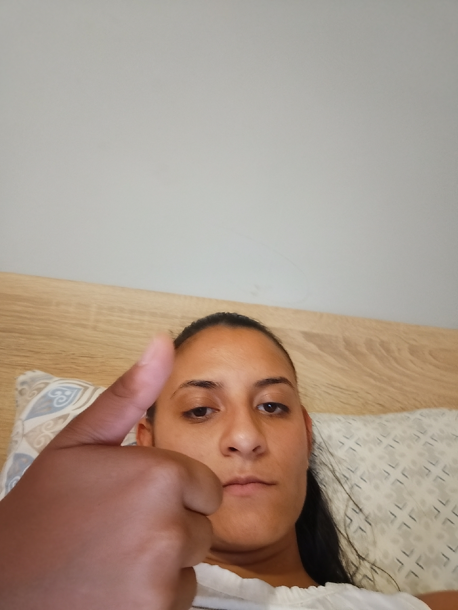 A woman is lying in bed, her head on a pillow, and gives a thumbs up to the camera. She has dark hair, dark eyes, and is wearing a white top. The background shows a wooden headboard and a grey wall. The scene appears to be indoors, likely in a bedroom. There is no specific information that can identify the location as Yambol, Bulgaria.