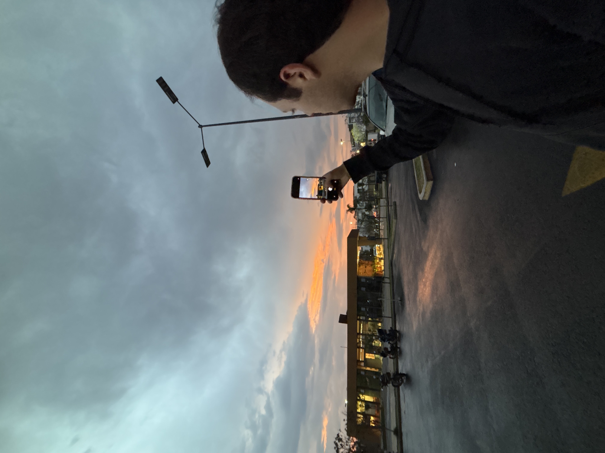 A man stands at the edge of a wet parking lot, holding a smartphone in his right hand to capture a photo of the sky. The sky is divided between a cloudy blue-gray and a vibrant orange sunset. In the background, a building with illuminated windows is visible, suggesting a commercial establishment. A lamppost stands near the man. The city is Santa Clara Ocoyucan, Mexico.