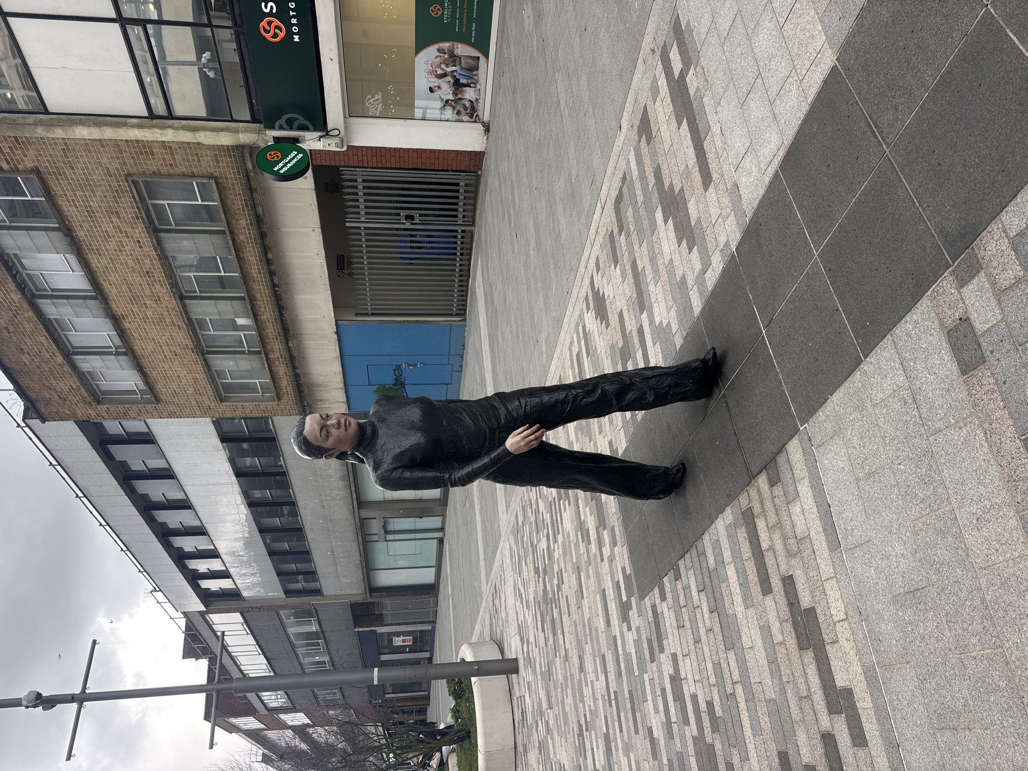 The image captures a life-sized, dark-colored statue of a person in mid-stride, positioned on a pedestrianized street in Woking, United Kingdom. The statue wears a high-collared, long-sleeved top and wide-leg trousers, all appearing as a cohesive, textured dark garment. Its hair is pulled back, and the face presents a neutral expression.
The ground comprises a paved surface featuring rectangular paving stones in various shades of grey and pinkish-grey, arranged in patterns. A distinct strip of darker grey paving runs directly beneath the statue. To the left, a tall, dark grey lamppost stands beside a low concrete planter containing a bare tree or shrub.
In the background, a row of multi-story commercial buildings lines the street under an overcast sky. Buildings on the left exhibit brick facades with numerous windows, some appearing vacant. On the right, a building displays a green circular sign marked "MORTGAGES INSURANCES" with an 'S' logo. Below this, a storefront window features an advertisement for "STERLING STREET" with an image of people on a sofa. Adjacent to the storefront is a metal-barred gate and a blue boarded section displaying graffiti. Small birds are faintly visible in the sky above the buildings. Minimal pedestrian presence is noted in the distant background on the left.