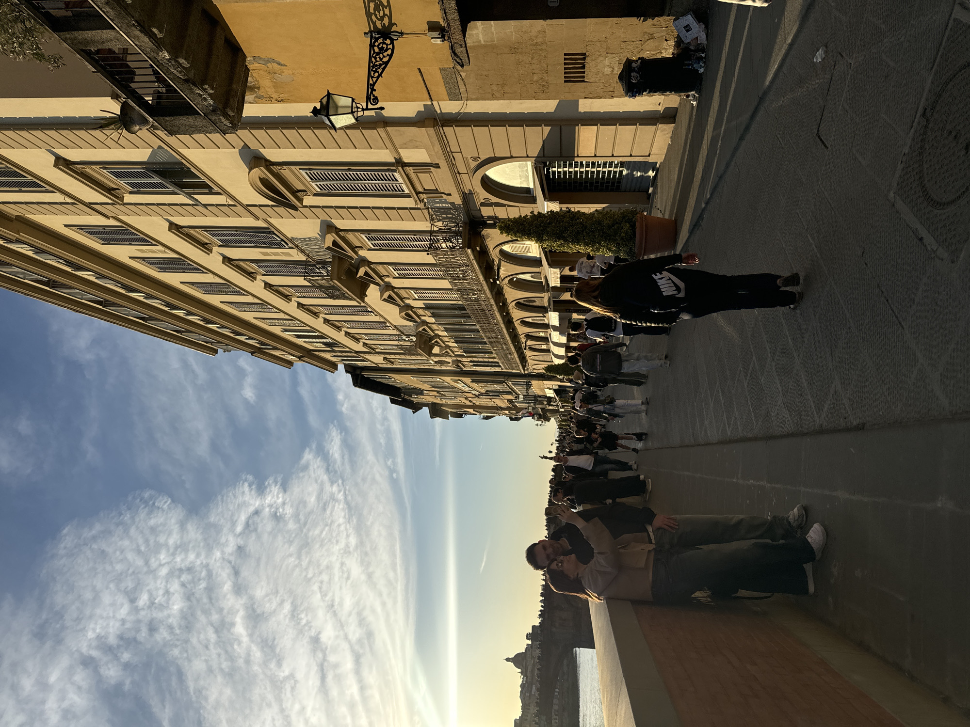 The image shows a street scene in Florence, Italy. The view is taken from a bridge, likely Ponte Vecchio, with the Florence skyline visible in the background. In the foreground, a couple is sitting on the bridge's wall, looking towards the skyline. A large group of tourists is walking along the sidewalk beside the buildings. The architecture suggests a historic European street with multi-story buildings featuring rows of windows, some with shutters, and decorative architectural elements. The sky is partly cloudy with a warm glow, indicating either early morning or late afternoon. A street lamp is visible on the wall. The sidewalk is paved with dark stone tiles. A garbage container is seen close to the building.