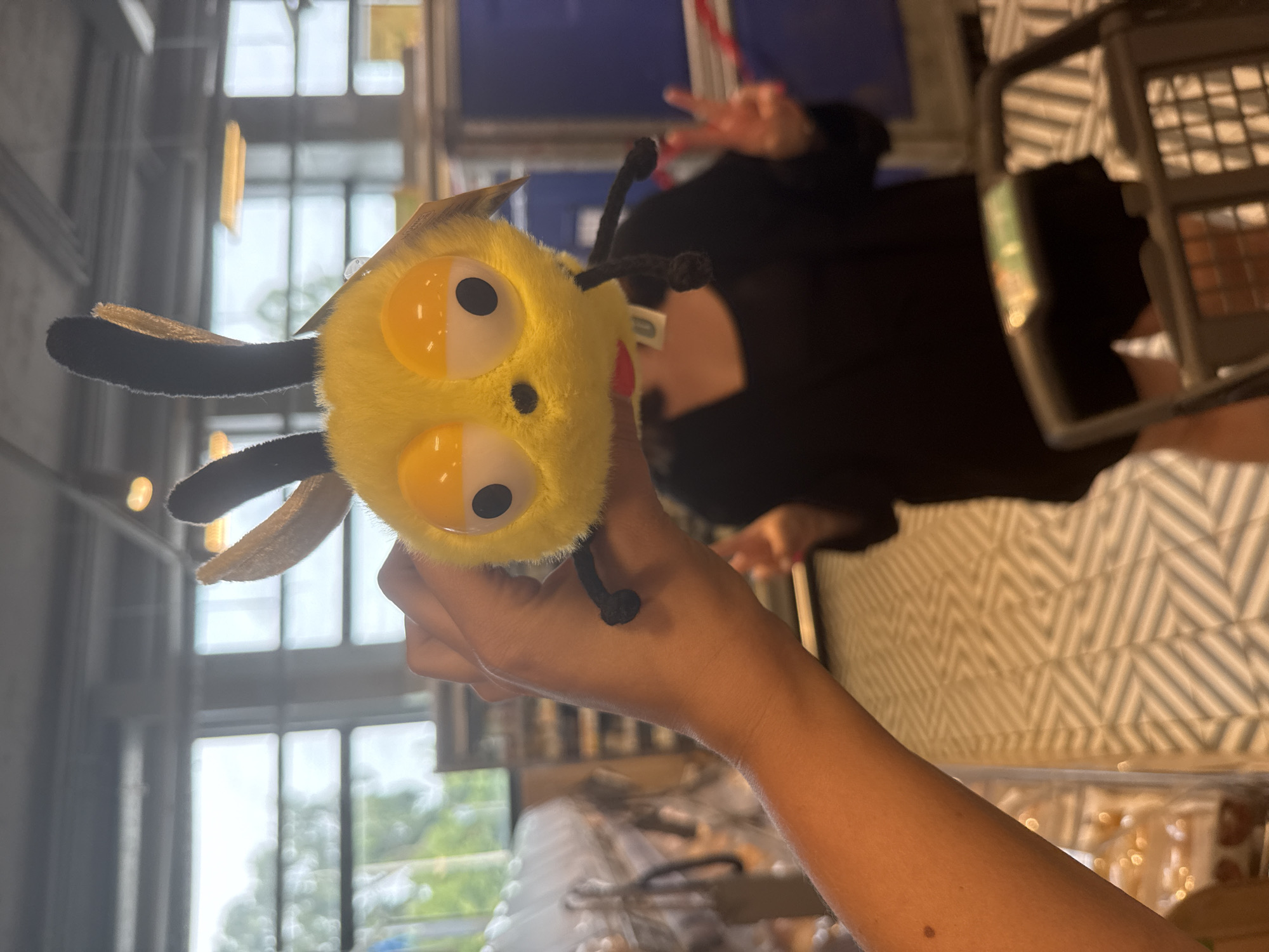 In this vertically oriented image, a plush toy bee is held in a person's hand. The bee is bright yellow with large, round eyes that are half white and half yellow, and small black pupils. It has black pipe cleaner antennae that are bent and fuzzy. Two fuzzy beige wings extend from the sides of its head. The background is blurred, but suggests an indoor setting with large windows, possibly a store or warehouse. Outside the windows, trees and greenery are visible, hinting at daylight.
Behind the bee and to the right, a person wearing a black top is partially visible, holding up two fingers in a peace sign. The person's arm and hand are in focus, as is the plush toy. To the far right of the image, a shopping cart is visible, with its wire basket and handle. The floor has a patterned surface with diagonal stripes in white and a lighter neutral color. The overall impression is a casual snapshot taken during the daytime, likely in a retail or public space. The plush toy is the central focus, and its cheerful appearance may suggest a playful or happy mood. No discernible text is visible on the toy or in the background. The location context provided is Dmitrov, Russia.