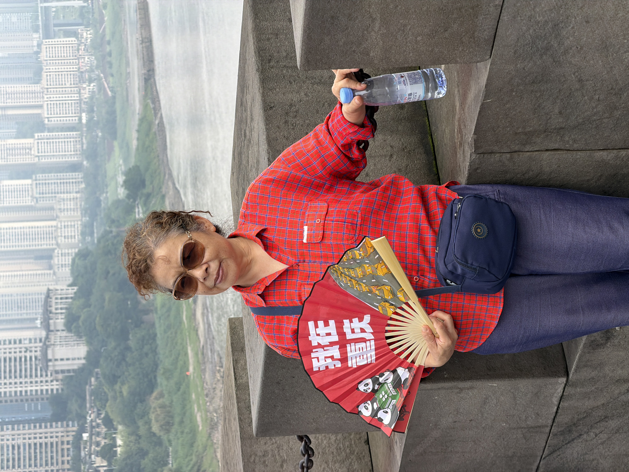 A woman stands on a stone ledge overlooking a city and a river. She is wearing a red and blue plaid shirt, blue pants, and sunglasses. She is holding a red fan with pandas and yellow cars depicted on it. In her right hand, she holds a clear plastic bottle of water with a blue cap. The background shows a hazy cityscape with tall buildings and a bridge over a body of water. The environment appears to be outdoors on a cloudy day. The text on the fan appears to be Chinese characters. The overall scene is calm and depicts a tourist observing the city. The location is Yuzhong District, China.