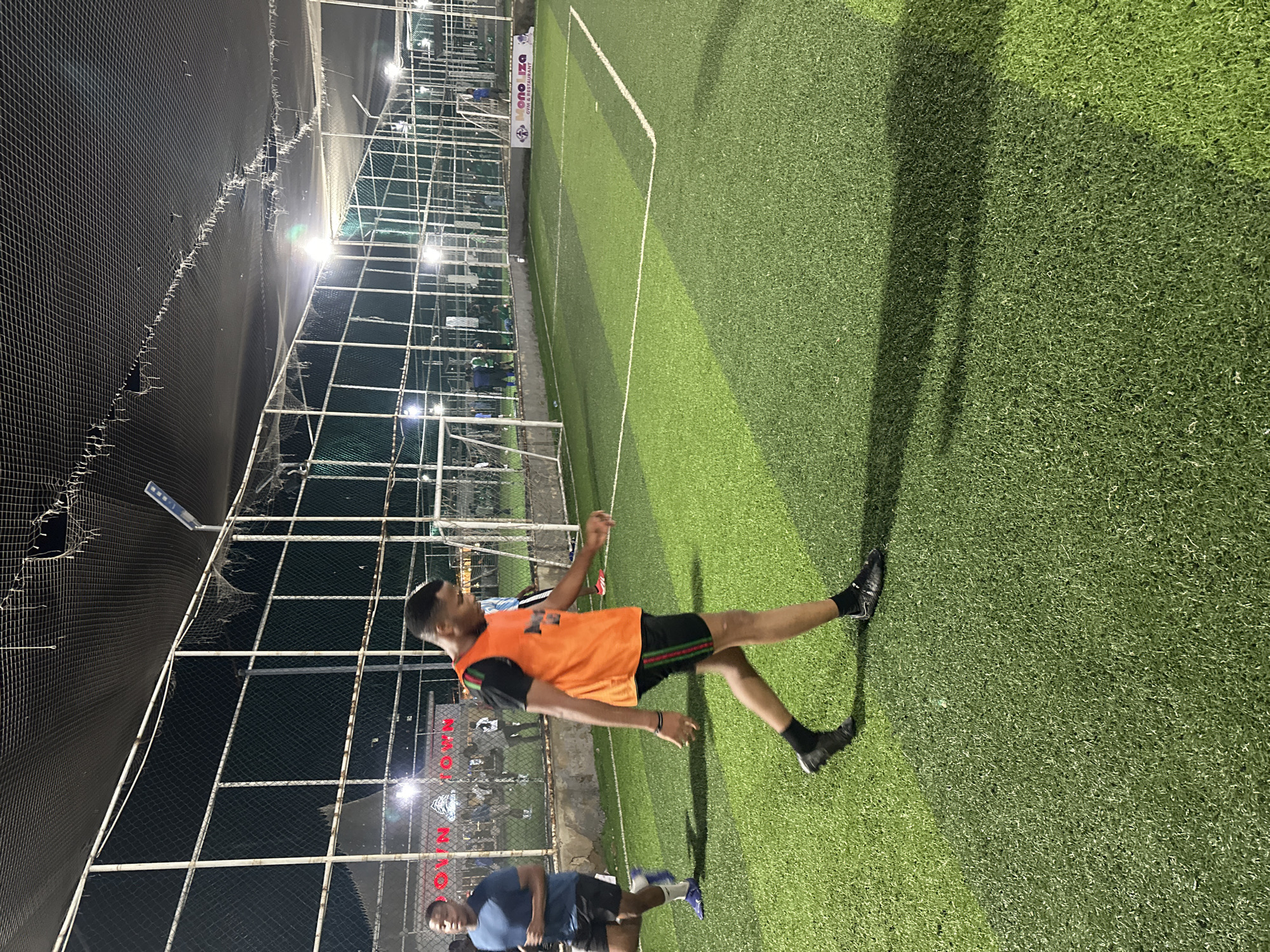 The image captures a night scene at a football pitch in Abuja, Nigeria. Two young Black men are present on the artificial turf field. One man, in the foreground and facing right, wears an orange training bib over a black t-shirt and black shorts with green and red stripes. He is walking. The other man, in the background and on the left, is wearing a blue t-shirt and black shorts, and appears to be running. The football pitch is well-lit by artificial lights mounted on poles, and a protective net covers the top of the enclosure. A fence surrounds the field, beyond which some buildings and more lights are visible. A sign with the text "MONO LIZA GYM & RESTAURANT" is visible on the right side of the field. The overall environment suggests an organized sports facility.