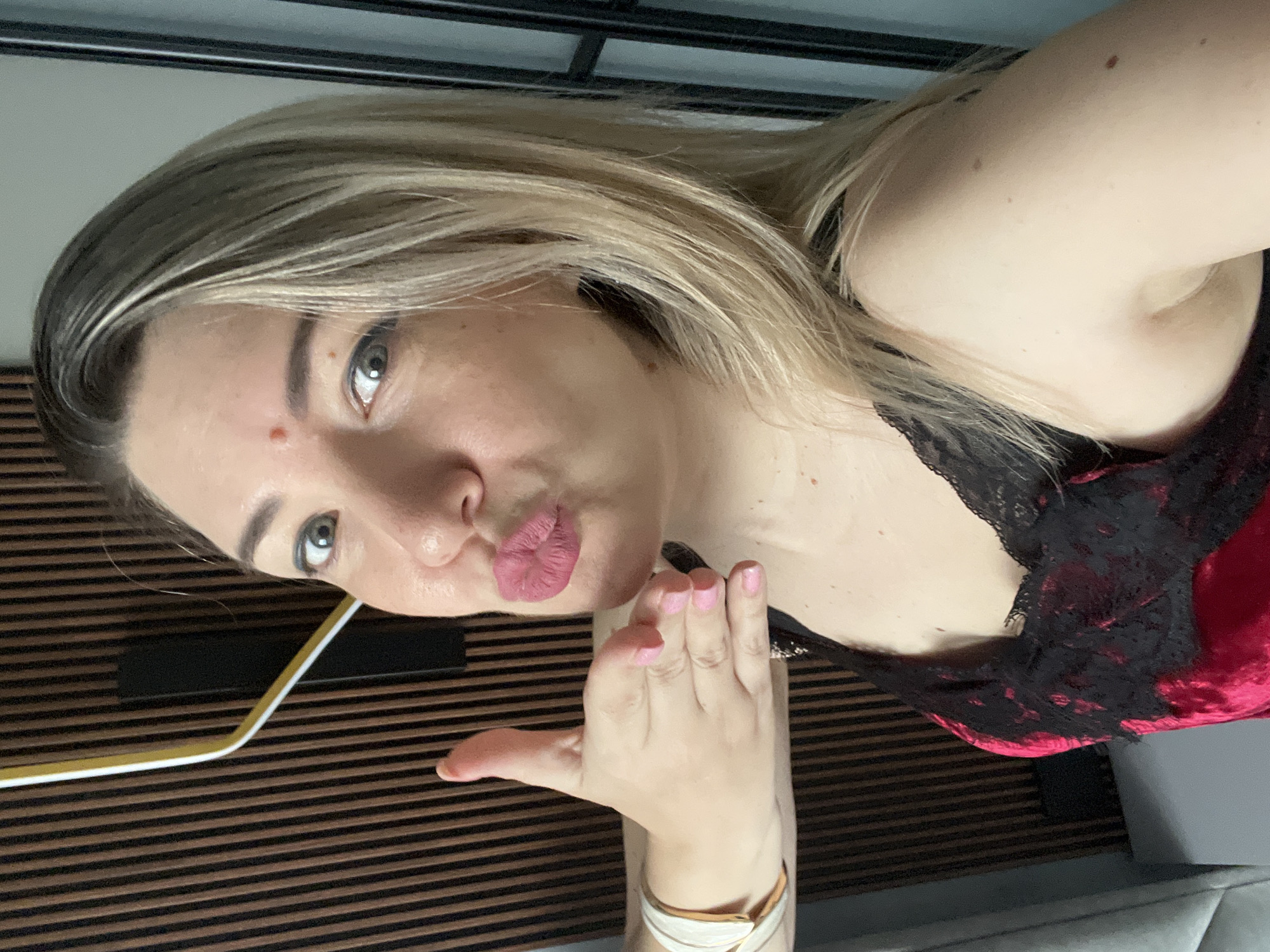 The media file is a portrait of a woman taken from a slightly high angle. She has blonde hair, which is styled in soft waves, and her eyes are a striking blue-gray. She is wearing a red velvet top with black lace trim. Her lips are pursed, and she has pink nail polish on her fingers, one of which is raised towards her face as if in a gesture of playful secrecy. She has a small mole on her left cheekbone and a few other small moles visible on her arm. The background consists of a striped wooden surface, suggesting a wall or furniture. A yellow and white object, possibly a straw or a pen, is visible on the left side of the frame, resting against the striped surface. The lighting appears to be indoor, soft, and diffused. The woman's expression is one of playful intimacy, with a hint of a smile. There is no visible text in the image. The setting, combined with the pose and attire, suggests a casual and personal moment captured.