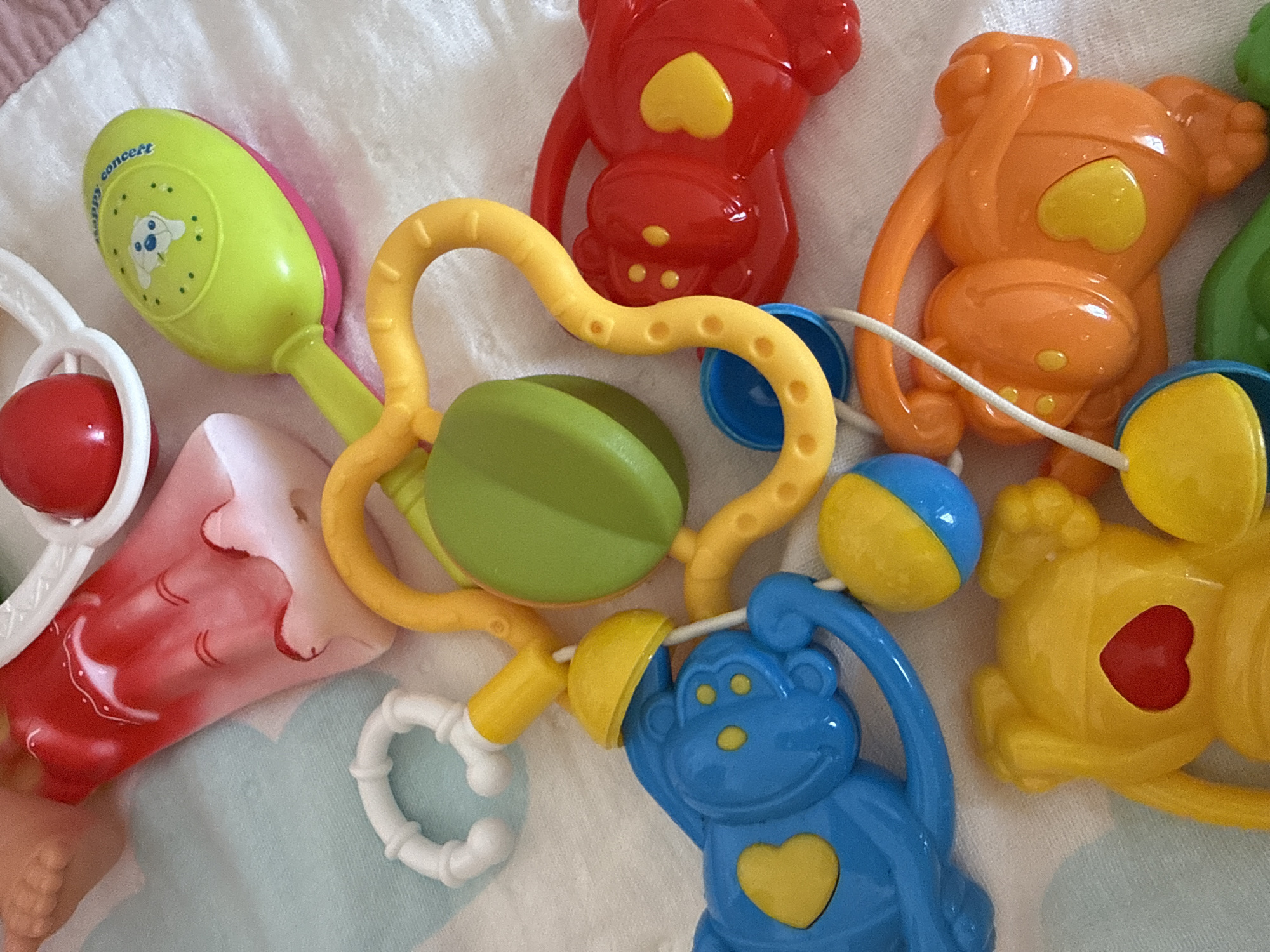 The image shows a collection of colorful plastic baby toys on a soft, patterned surface. The toys include animal shapes (bears and a monkey), rattles, and a teething ring. The toys are red, orange, yellow, green, and blue. The background is a soft, light-colored fabric with a subtle pattern.