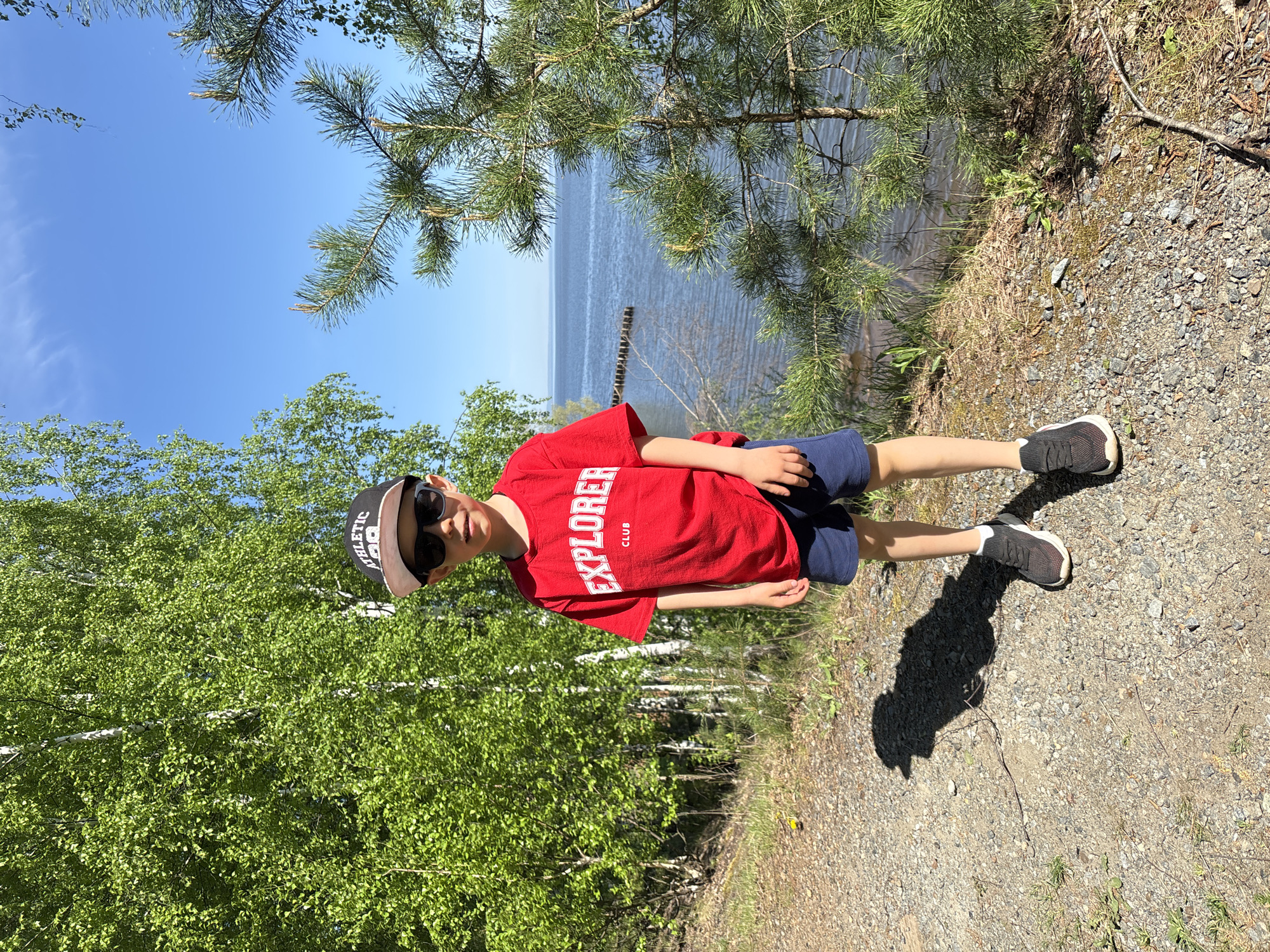 A young boy stands on a gravel path alongside a body of water on a sunny day in Chaikovsky, Russia. He wears a red t-shirt with "EXPLORER CLUB" printed on it, navy blue shorts, black and white athletic shoes, a baseball cap, and sunglasses. He is positioned between a birch tree forest on his left and the water on his right, which is visible through a pine tree branch hanging overhead. In the background, a clear blue sky stretches above the lake. Wooden poles are visible in the water near the shore. The boy's shadow falls on the gravel path.