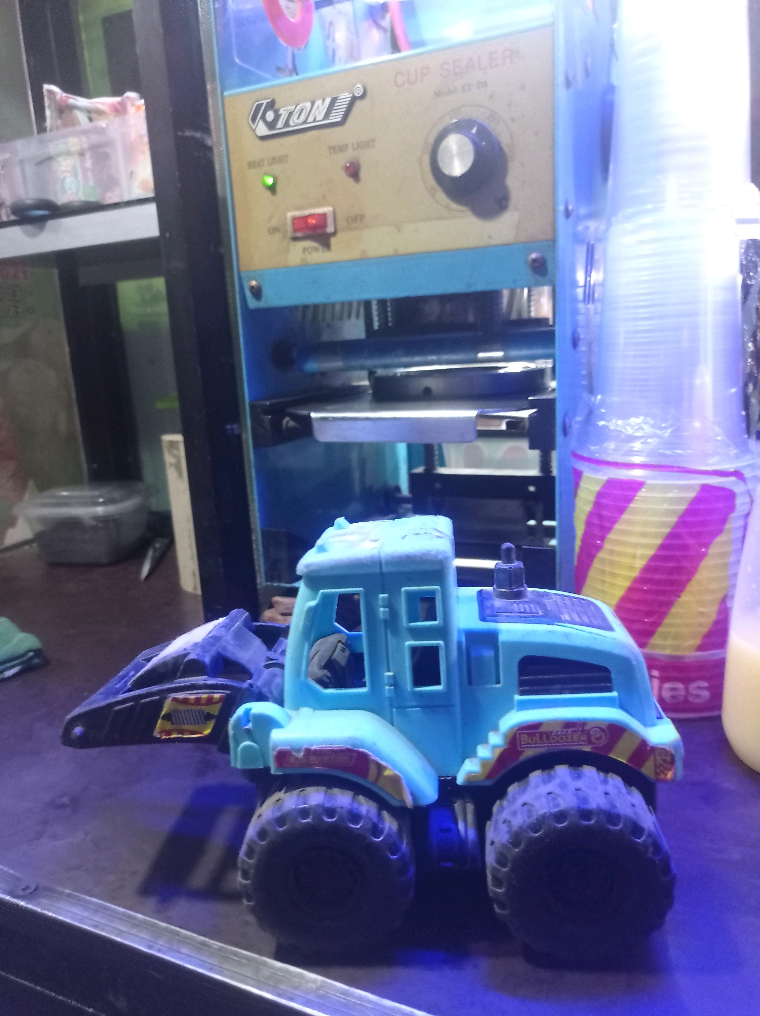 A blue toy truck with large black tires sits on a dark purple surface. The truck has a yellow and red stripe design on its sides and a black scoop at the front. Behind the toy truck is a cup sealing machine, blue and beige, with a red power switch and two indicator lights, one green and one red. To the right of the cup sealer, a stack of translucent plastic cups with yellow and pink stripes is visible. In the background, a clear plastic container and other objects are partially visible on shelves. The scene appears to be set in a shop or stall in Tuban, Indonesia.