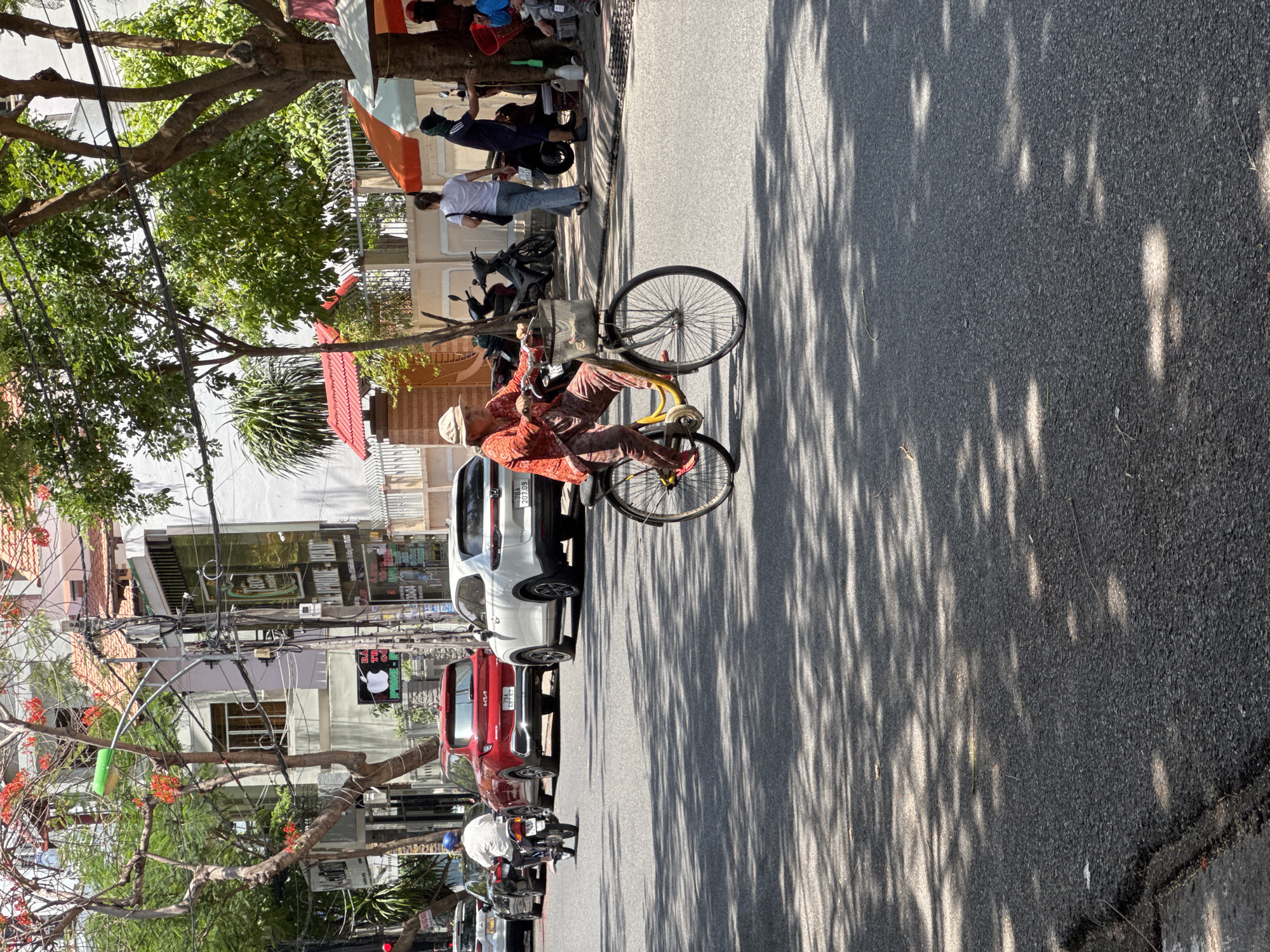 The image depicts a street scene in Nha Trang, Vietnam, on a sunny day. In the foreground, an elderly Vietnamese woman is riding a bicycle with a basket on the handlebars. She is wearing a light-colored hat, an orange patterned long-sleeved shirt, and patterned pants. The bicycle is yellow with black tires and spokes. The street is paved with asphalt and is dappled with sunlight filtering through tree shadows.
In the middle ground, a man is riding a motorcycle, wearing a white shirt and a blue helmet. Several cars are parked or driving on the street, including a red SUV and a white SUV. Buildings line the street, some with signs advertising businesses like "iPhone."
In the background, there are more buildings, trees, and utility poles with numerous wires. On the right side of the image, two people are standing under a colorful awning, and another person is visible further down the street. Motorbikes are parked along the sidewalk. The overall impression is of a busy urban street in Vietnam on a warm day.
